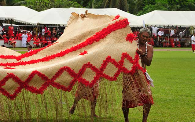  Samoan men carrying red and tan colored 'Ie Toga (Fine Mat) in ceremony