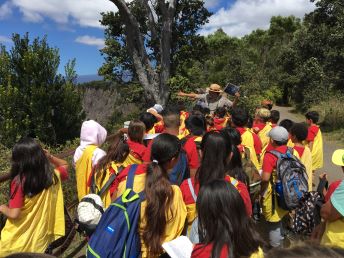 A park ranger in a straw flathat points towards a ledge to his right. A group of students look towards the ranger.