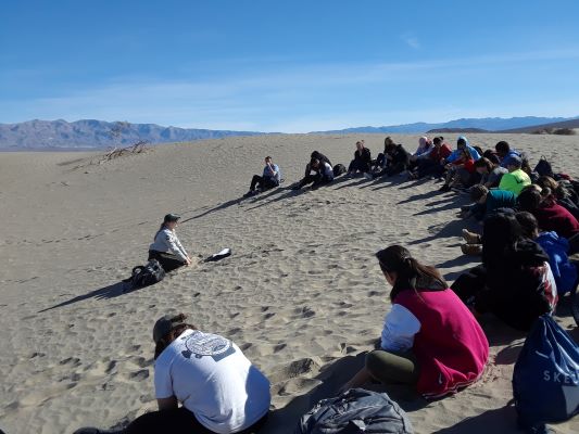 Students sitting in a half circle on the top of a sand dune face a ranger who is digging in the sand. Students sitting in a half circle on the top of a sand dune face a ranger who is digging in the sand.