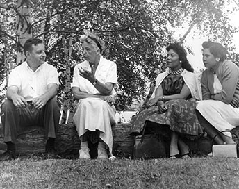 A man and three women sitting on a log outdoors in conversation. A man and three women sitting on a log outdoors in conversation.