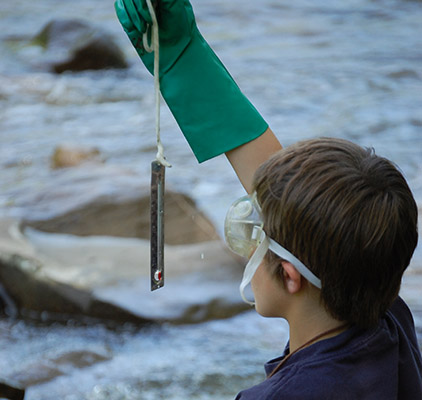 A child wearing safety goggles and a green rubber glove holds up a thermometer dangling from a string.