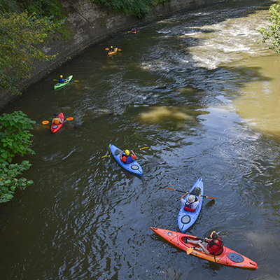 Aerial view of six brightly colored kayaks making their way toward riffles in a brown river.
