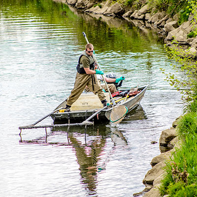 A man in beige waders and a life vest stands at the bow of a metal boat holding a long net with both hands.