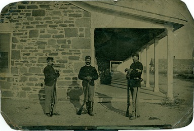 A black and white photo of three men in army uniforms standing in-front of a stone building.