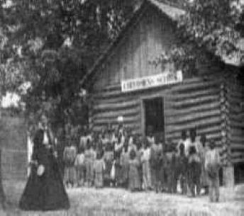 A white teacher in 19th Century clothes in front of her African American students in front of their log cabin school A white teacher in 19th Century clothes in front of her African American students in front of their log cabin school