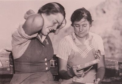 Elzada Clover and Lois Jotter sit next to each other looking at a book. Elzada Clover and Lois Jotter sit next to each other looking at a book.