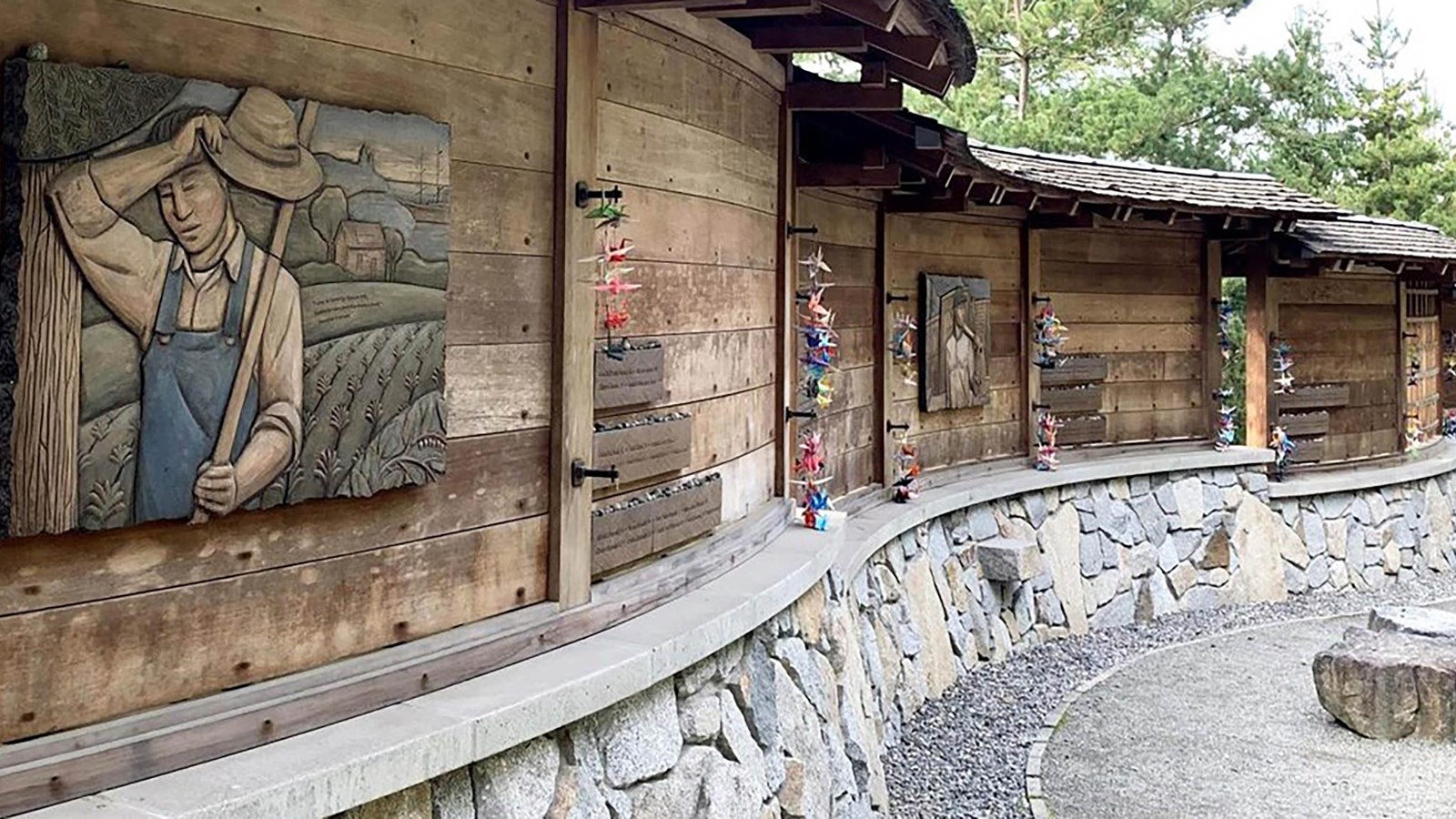 A curved wall of stone and wood with artistic depictions of people, and strings of paper cranes.
