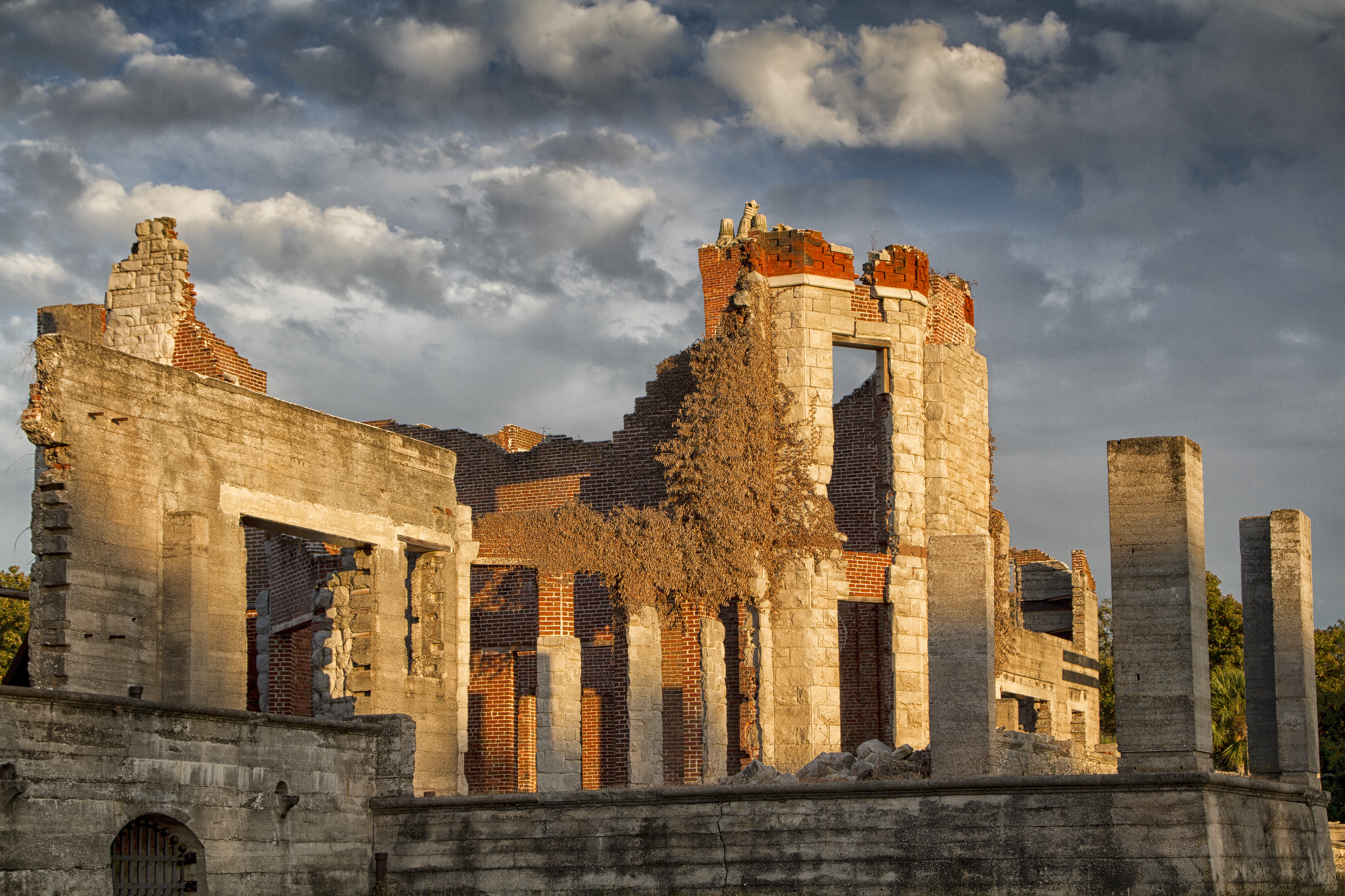 The brick and stonr ruins of a mansion are lit up by the evening sun with patchy clouds behind.