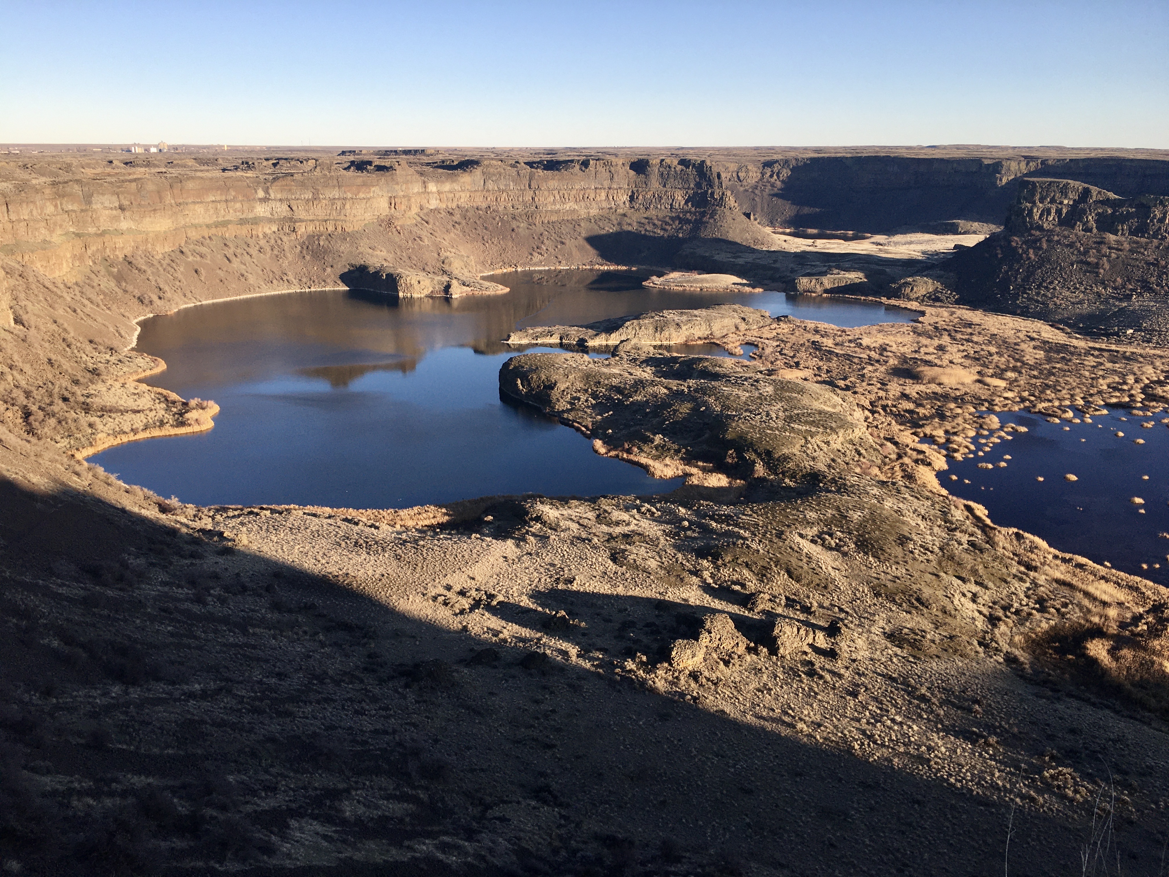 A broad amphitheater of tall cliffs of dark volcanic rocks towers over a series of small lakes.
