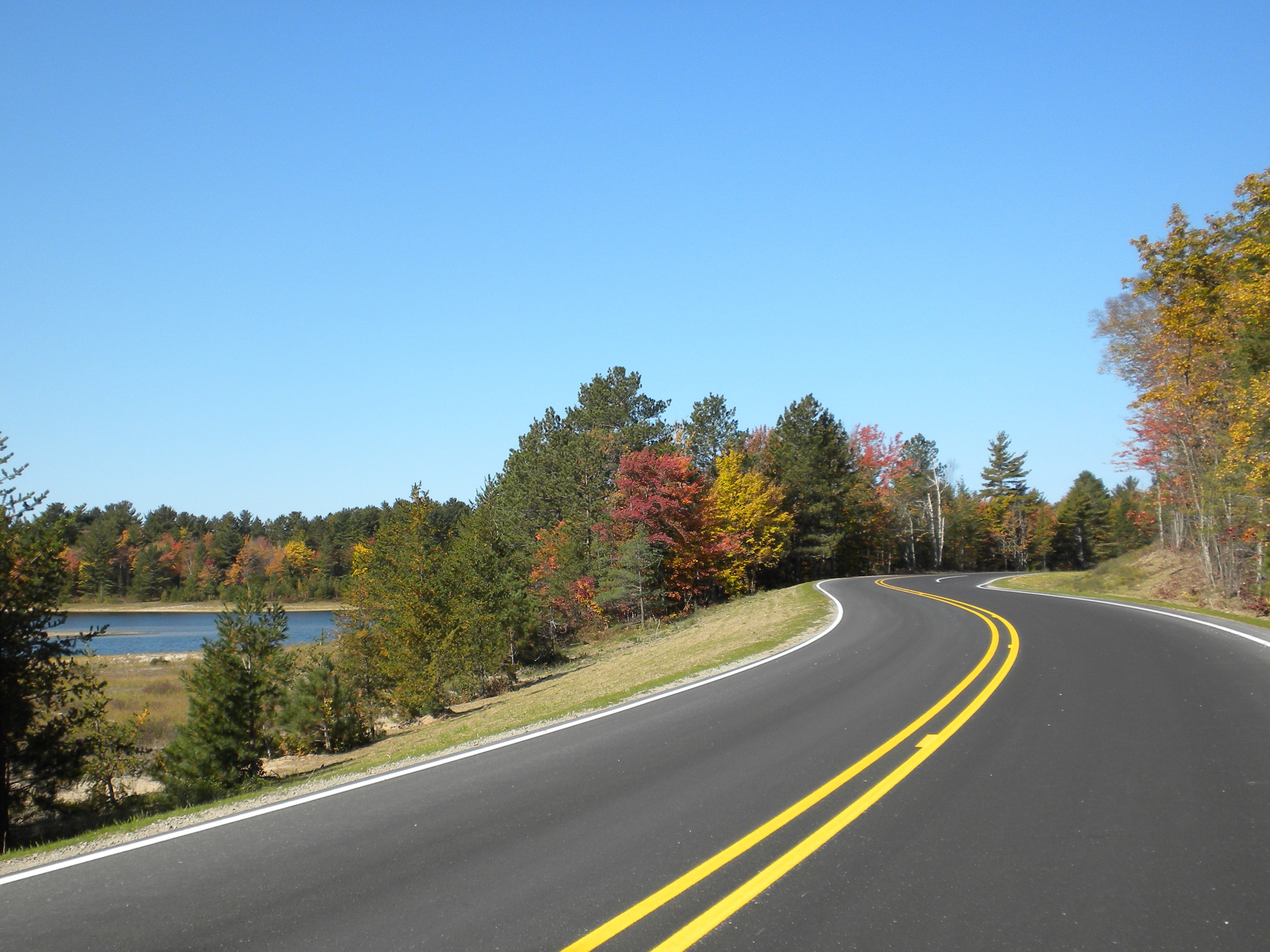 Paved county road through autumn forest with inland lake on the left.