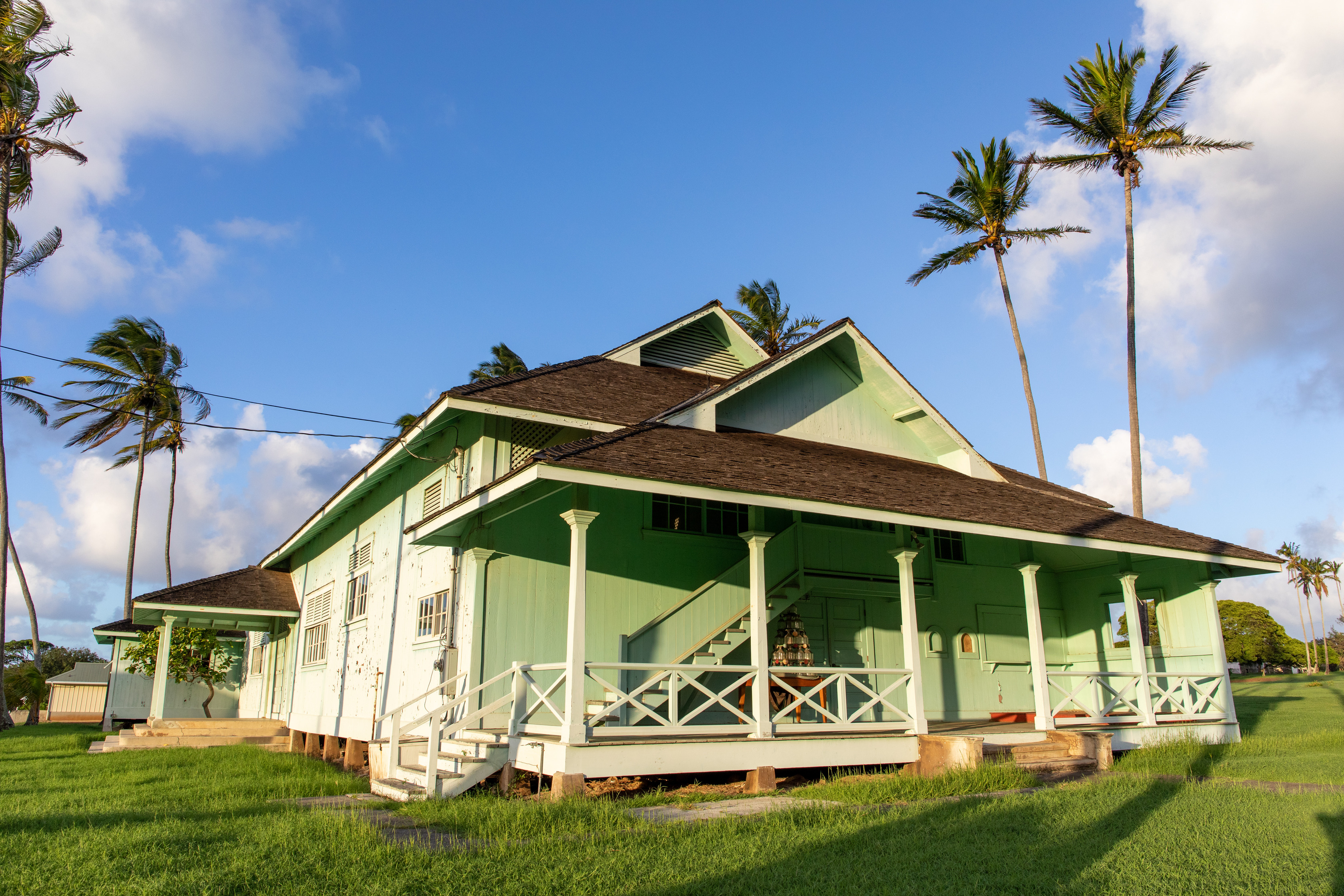 A large green building with a darker roof, surrounded by a lawn and palm trees.