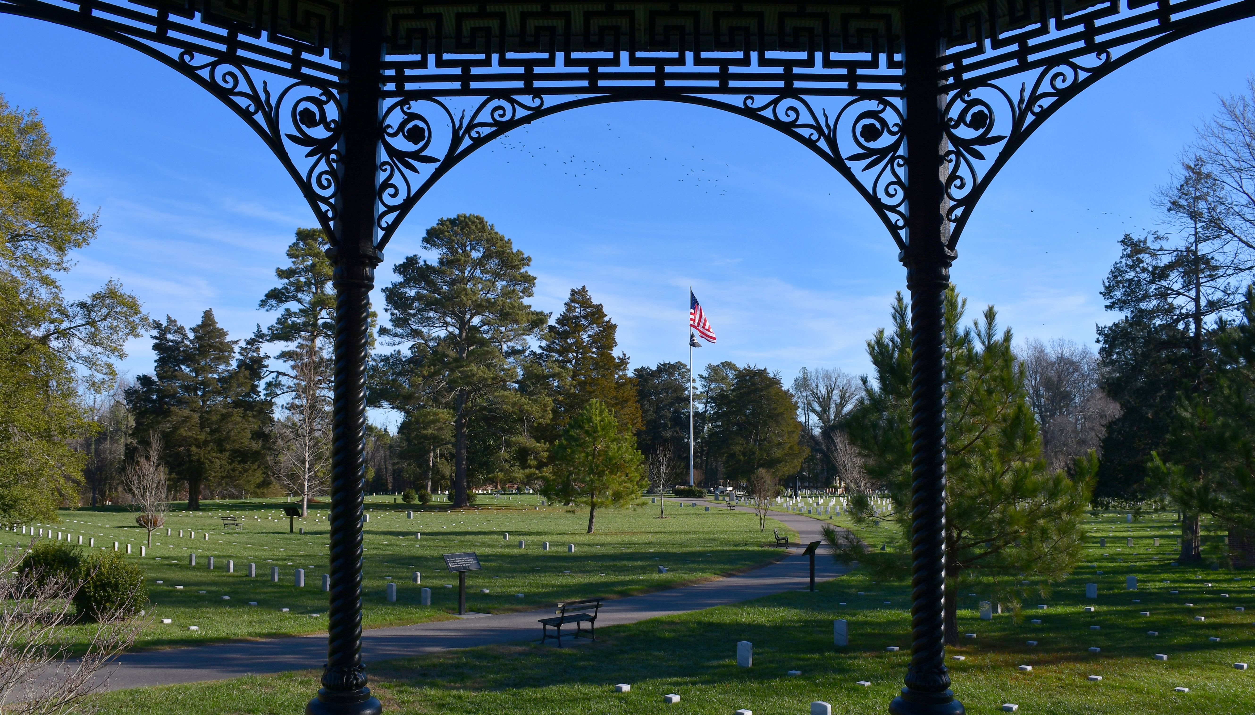 Two iron posts frame the American flag in the distance in the center of a cemetery on s sunny day.