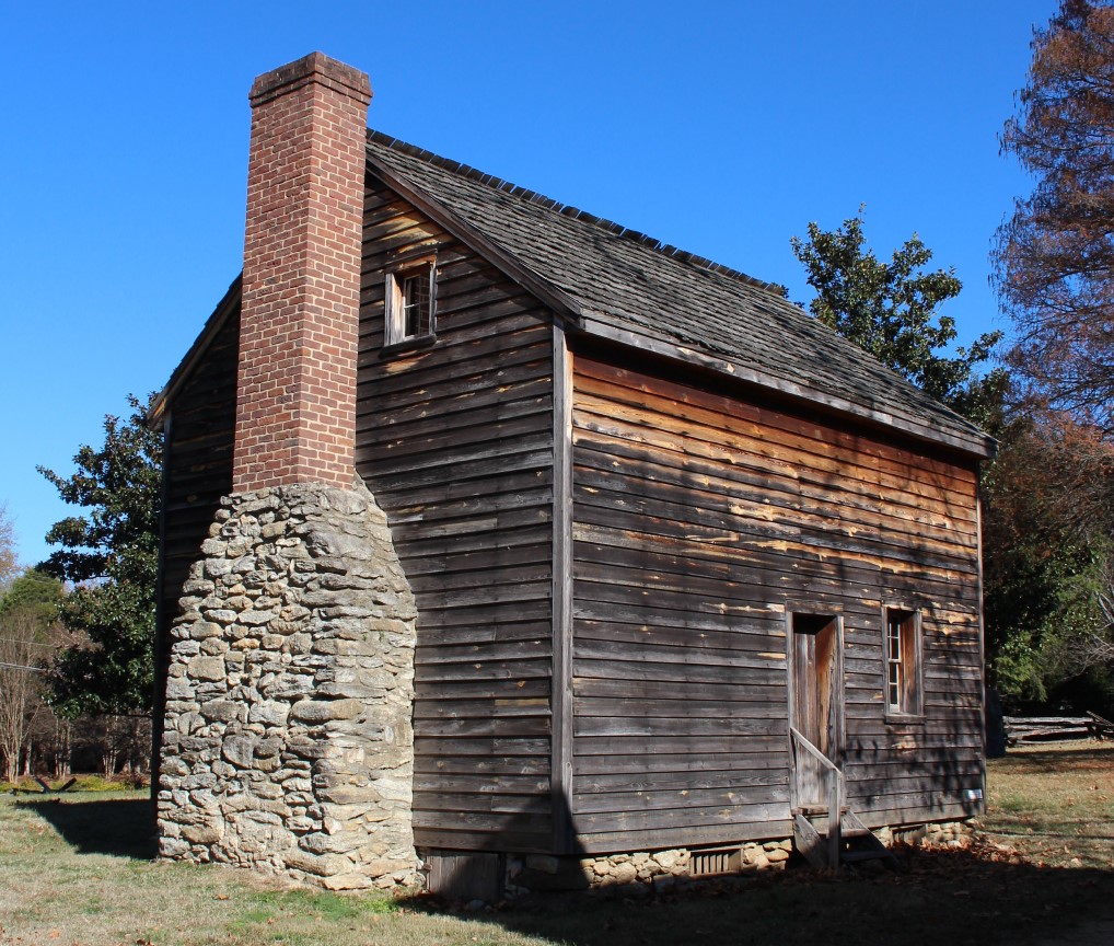 A two story building with clap board siding stands with trees close by. A chimney on its side.