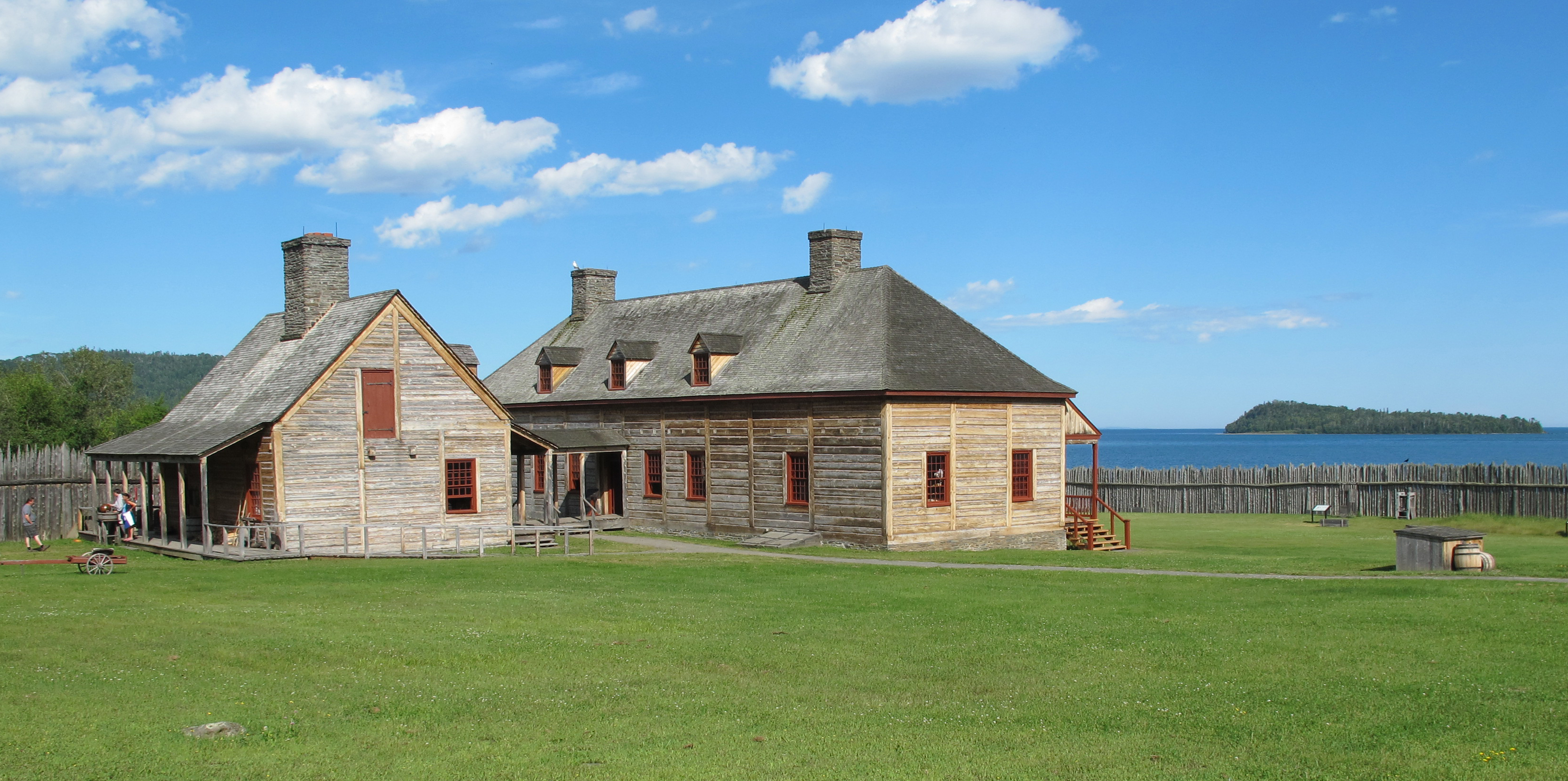 Historic wood buildings surrounded by a lawn with an island on a bay in the distance.