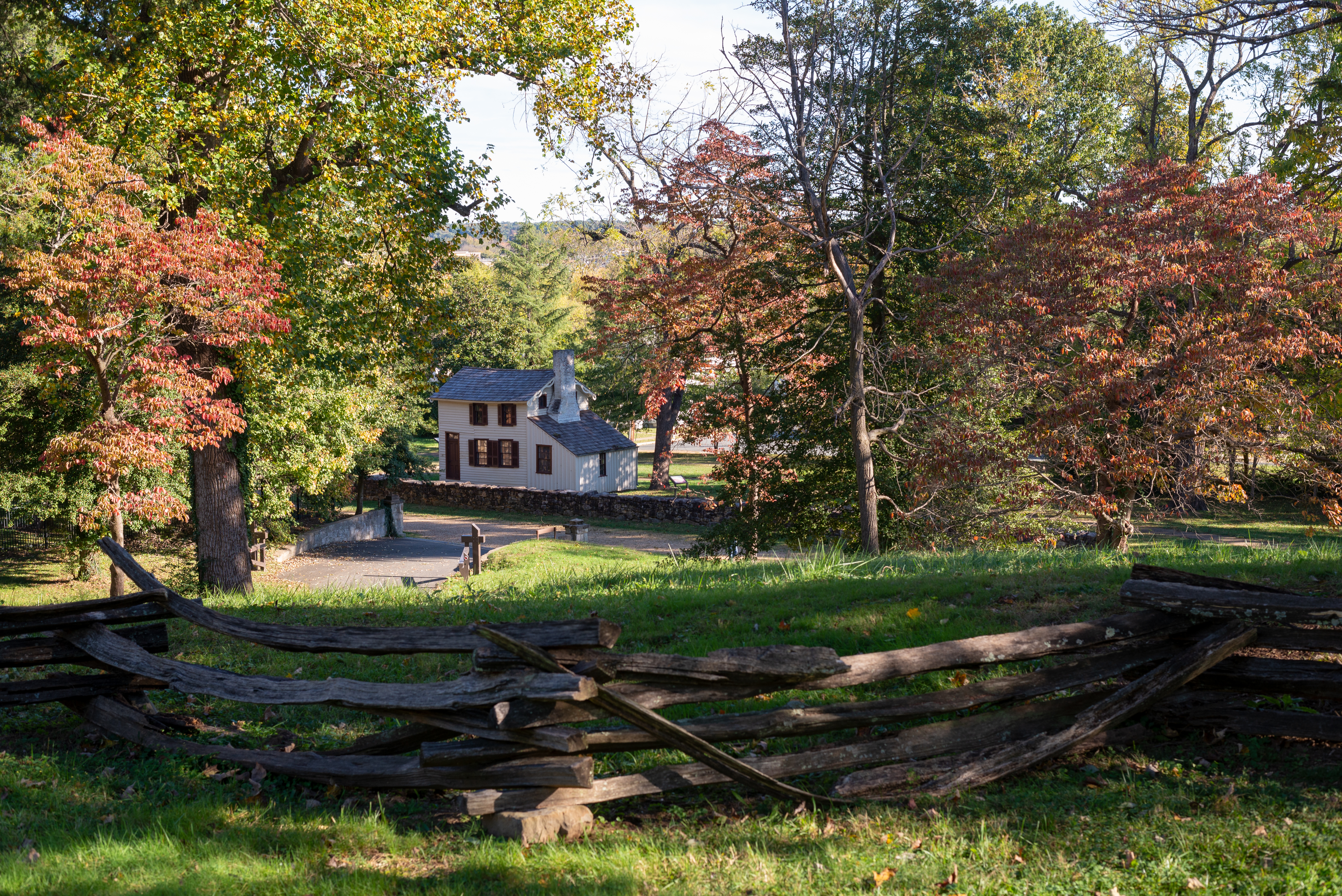 A view from atop a hill, through autumnal trees, to a gravel road with a small white house.