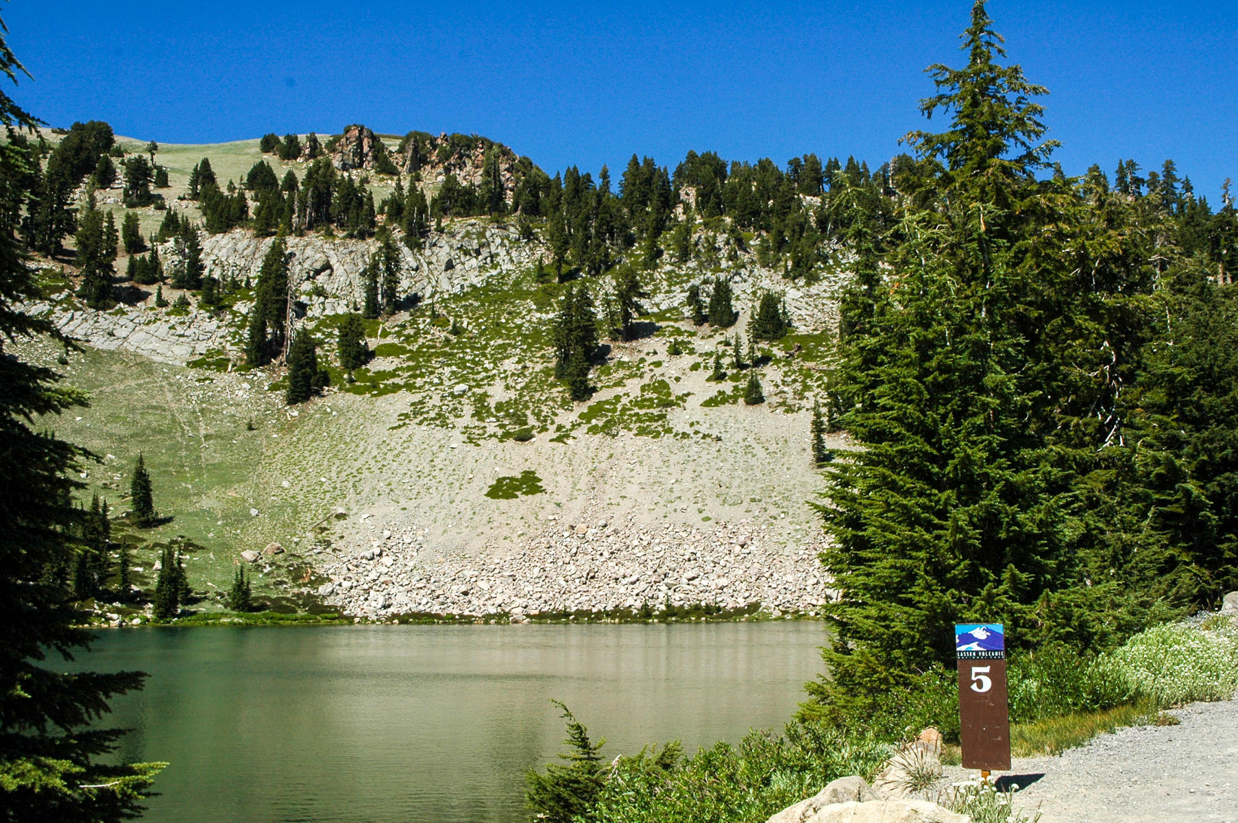 A photo of an alpine lake lined by conifers and steep rocky slopes with a small sign with "5."