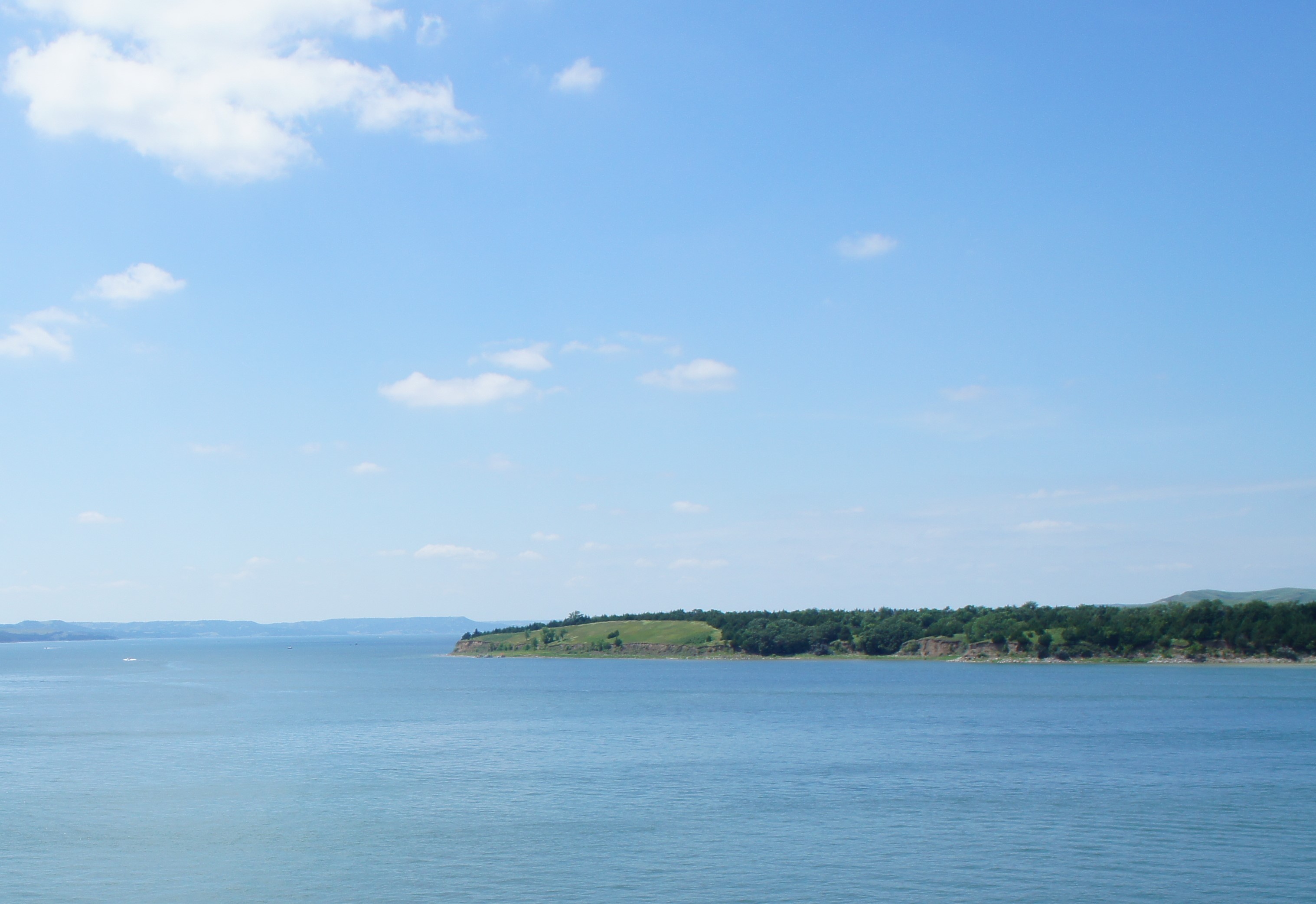 Missouri River with blue water and blue sky with sparse clouds. Green covered landform in foreground