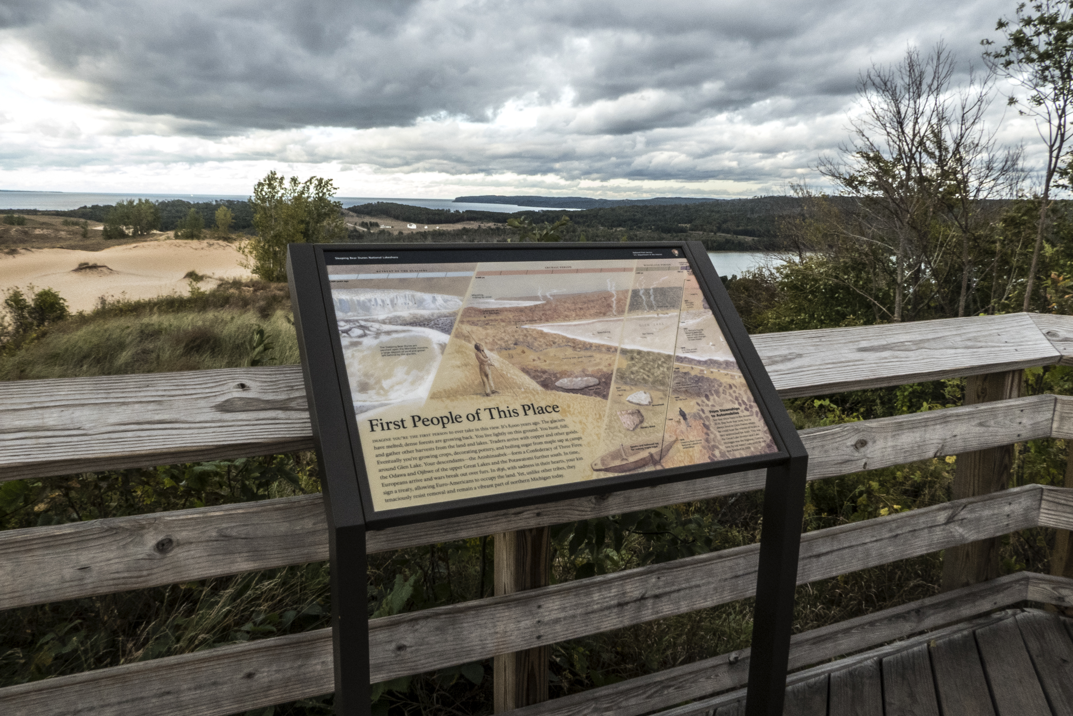A panel in a frame against a wooden railing with dunes, blue lake, and tree-covered ridges behind