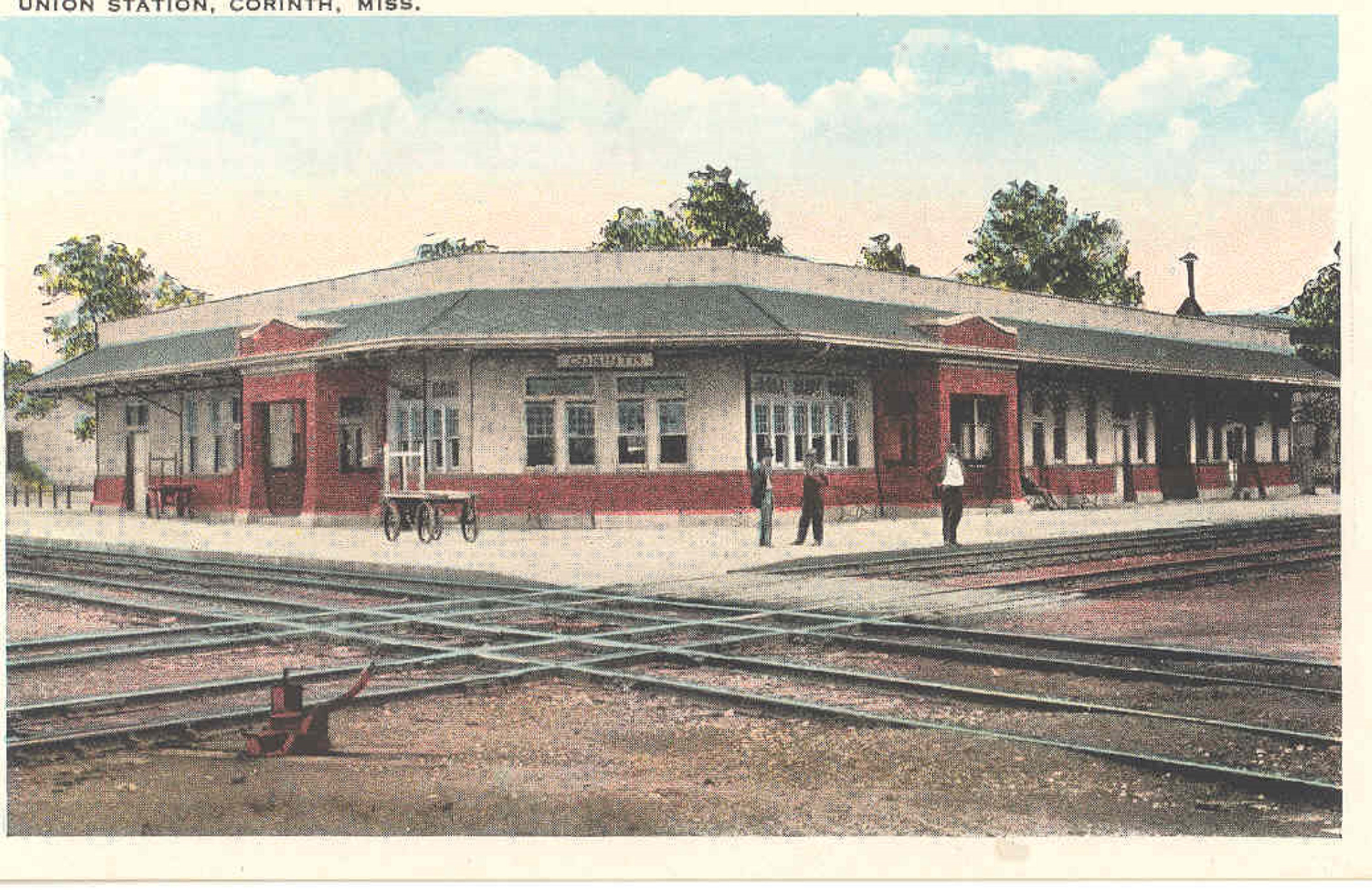 Two rail lines cross in the foreground, with a white and green building in the background.
