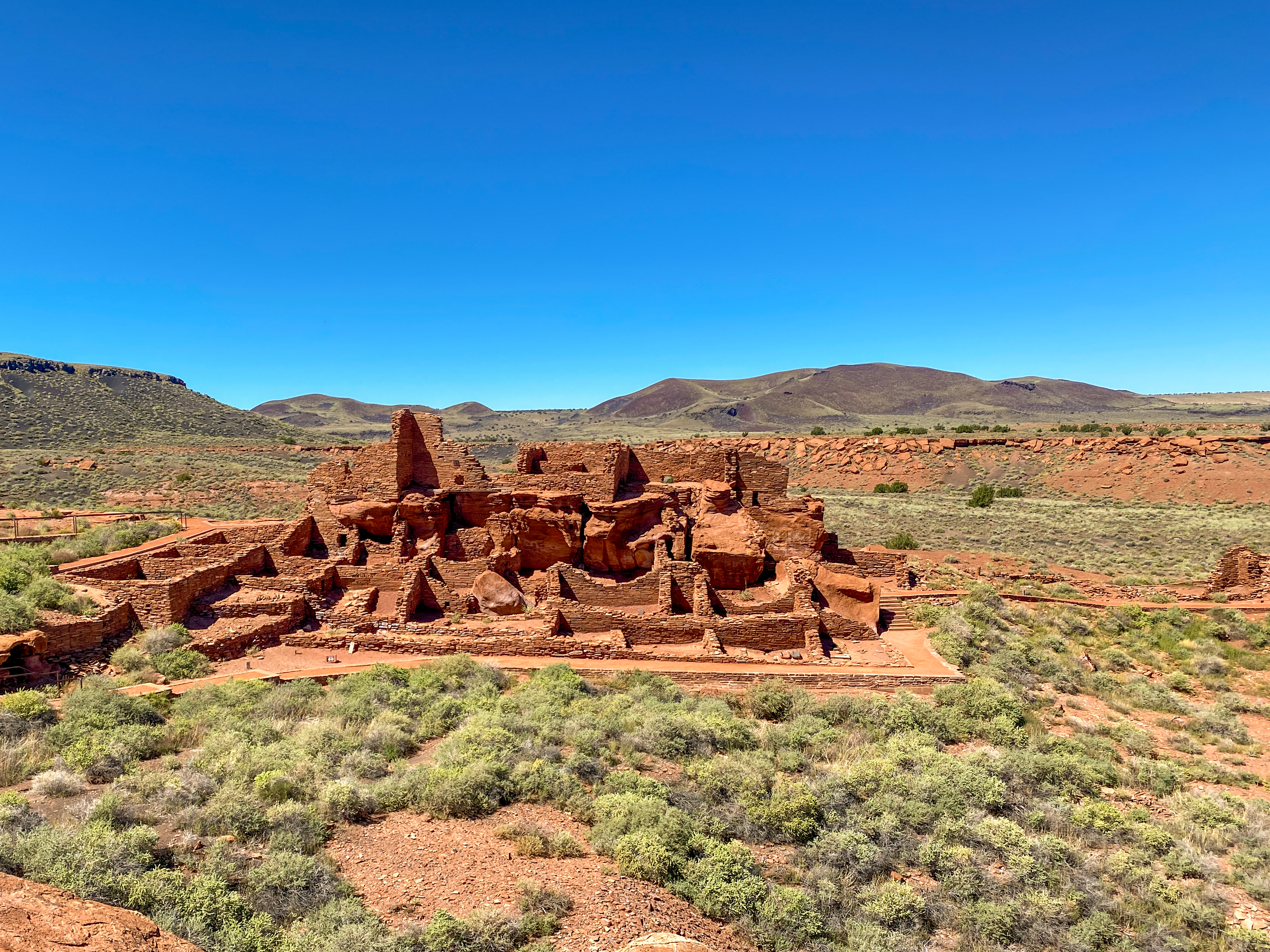 Blue skies above the desert landscape that holds a red rock pueblo with many rooms.