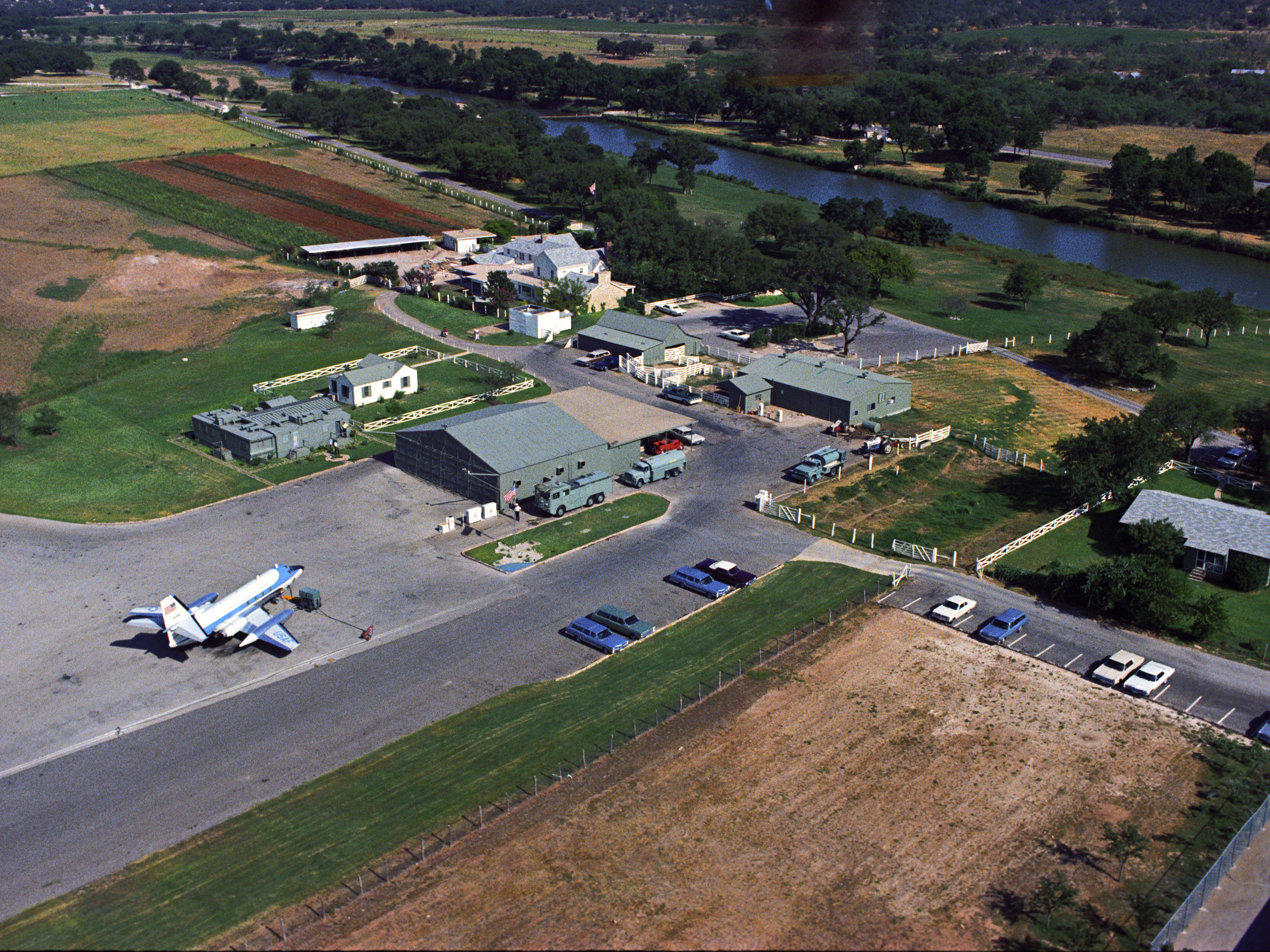 Aerial view of several buildings, 1960s era cars, and a jet surrounded by fields and river.