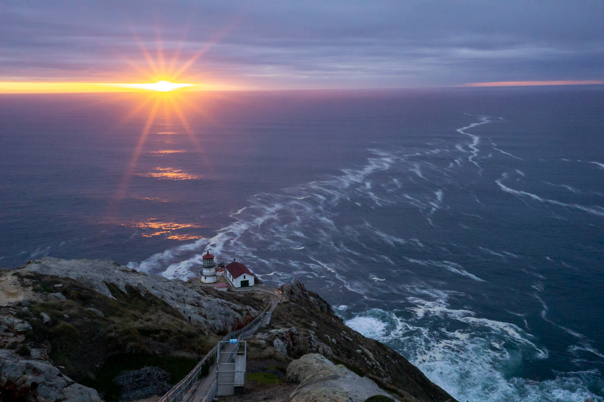 A short three story lighthouse on a rocky cliff overlooking the ocean at sunset.