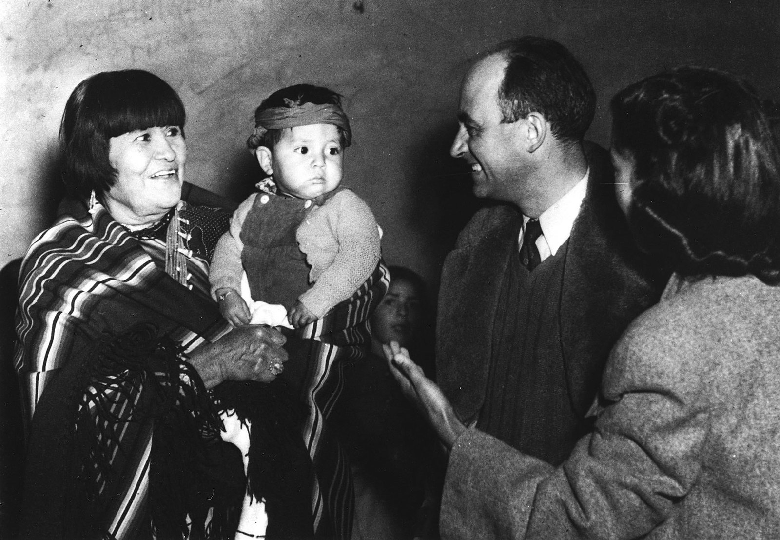 A black and white image of a Pueblo woman holding a baby in front of an older white couple.