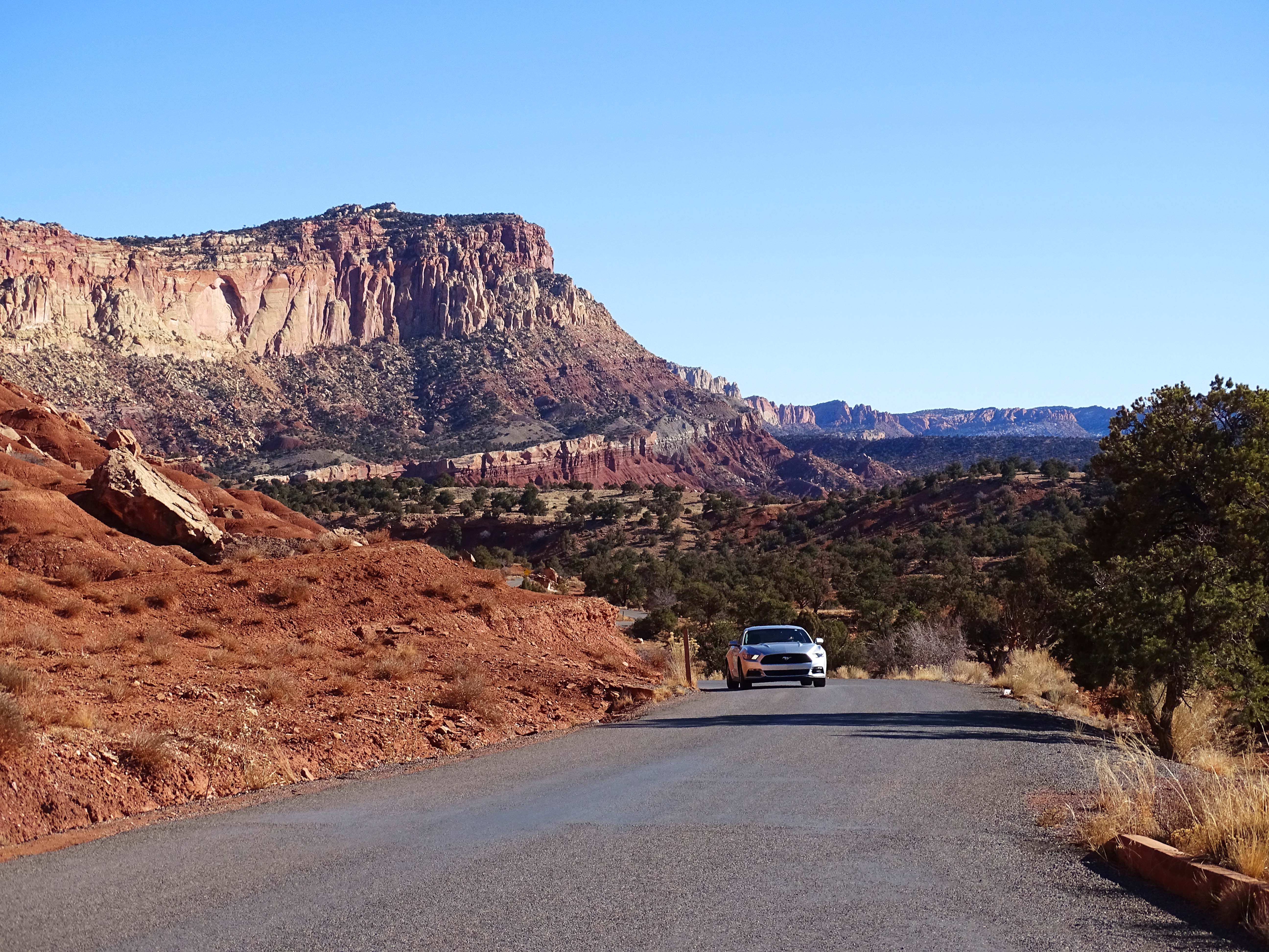 A silver car on a paved road next to huge banded cliffs of brown and tan.