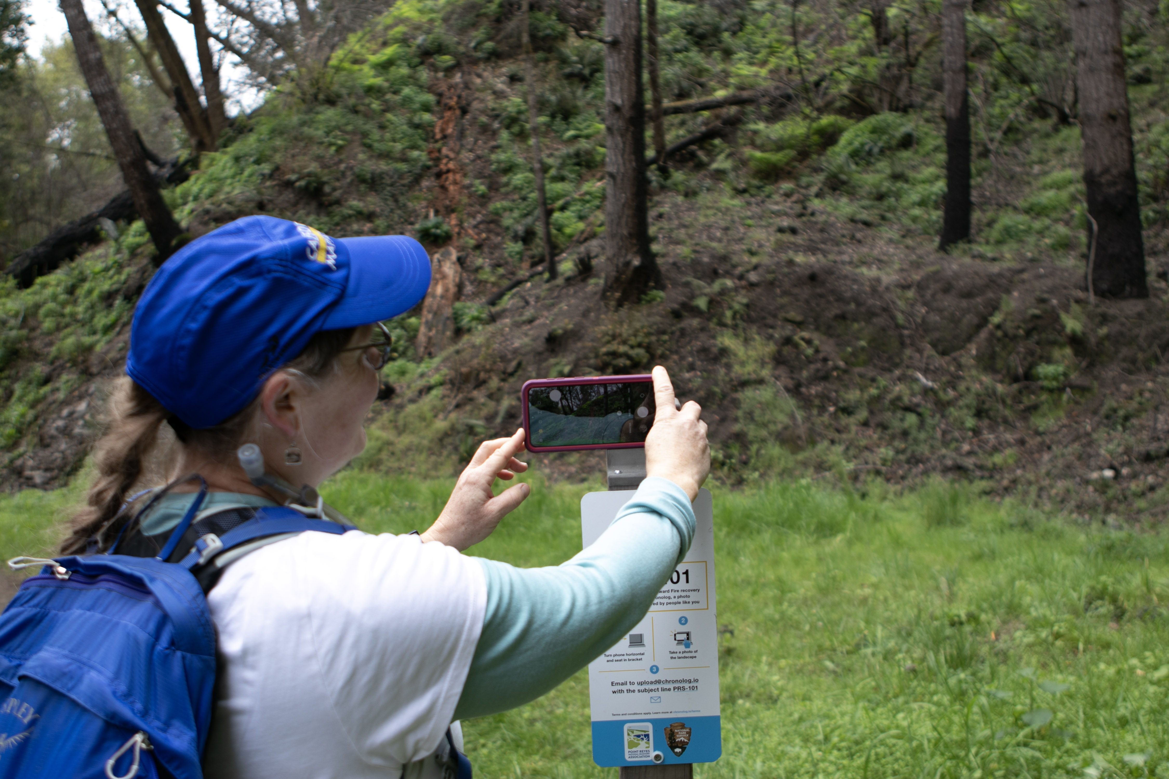 A woman takes a smartphone photo of a forested hill atop a bracket with a sign that reads "PRS 101"