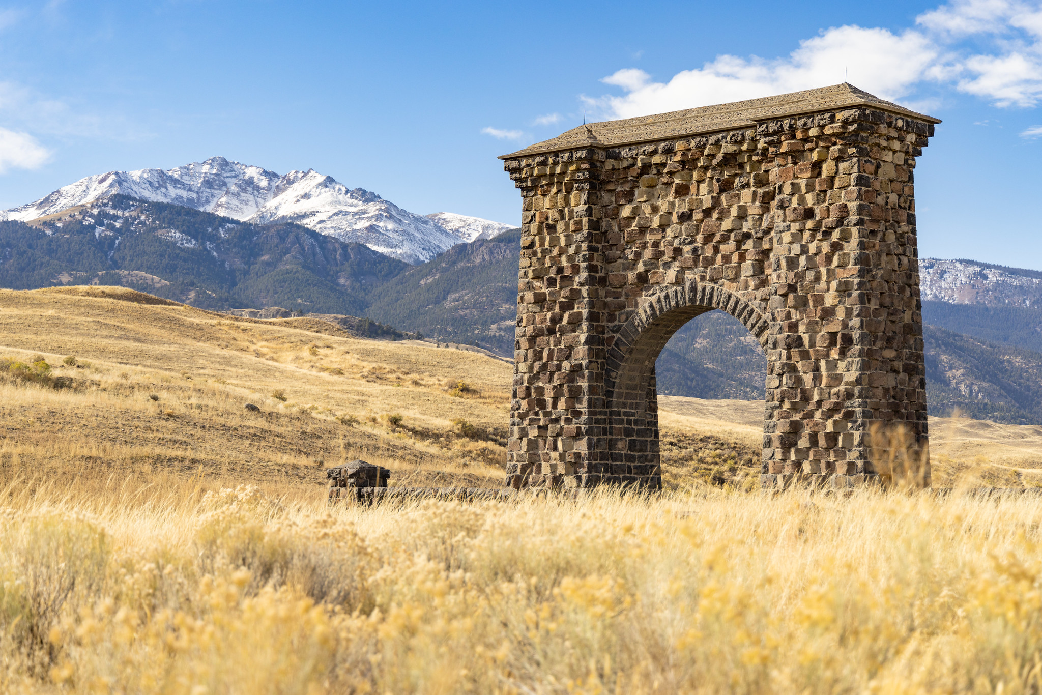A large stone archway, the Roosevelt Arch, stands in front of a snow covered mountain