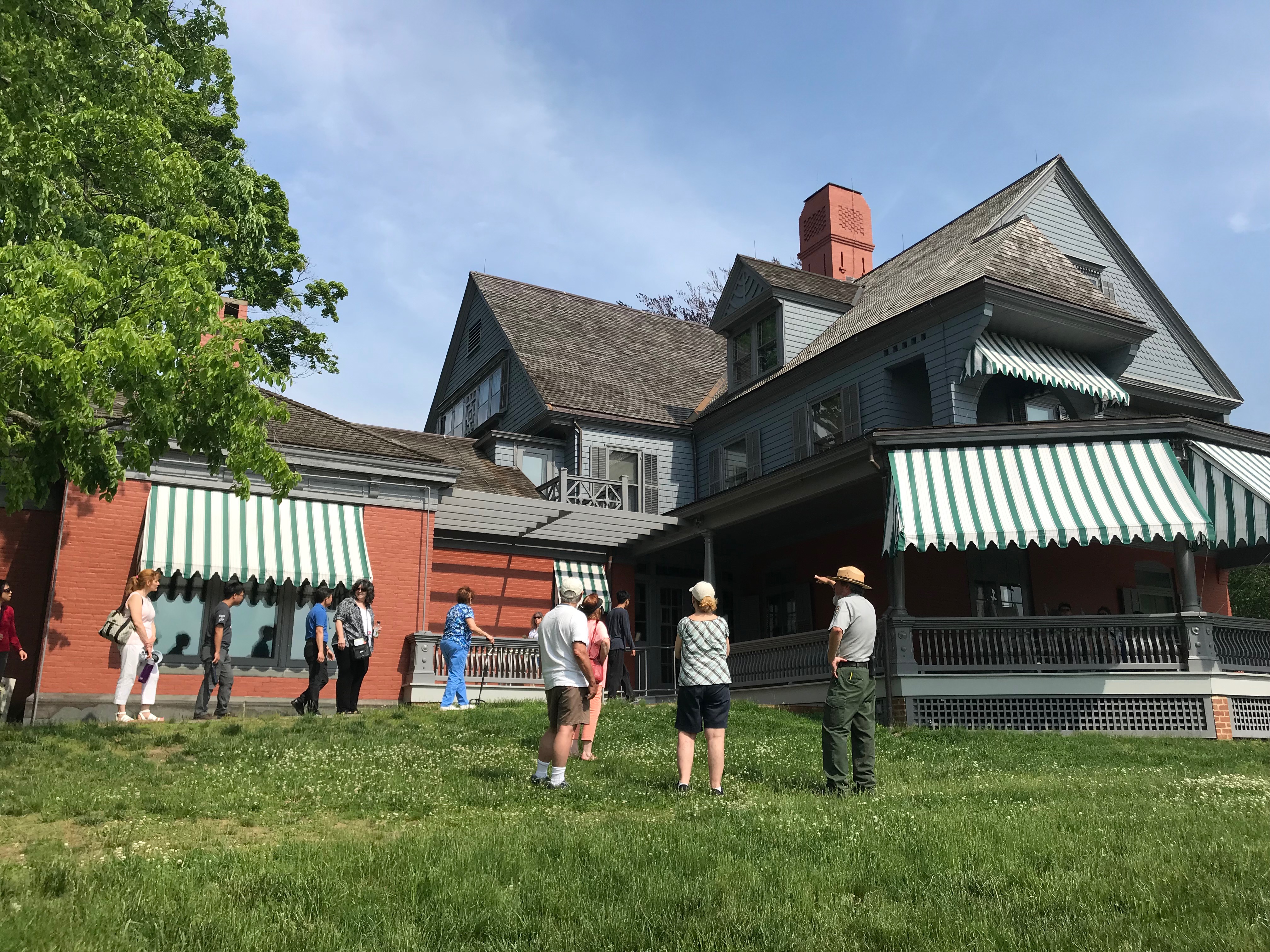 A park ranger points to a large gray and brick colored house with several visitors nearby.