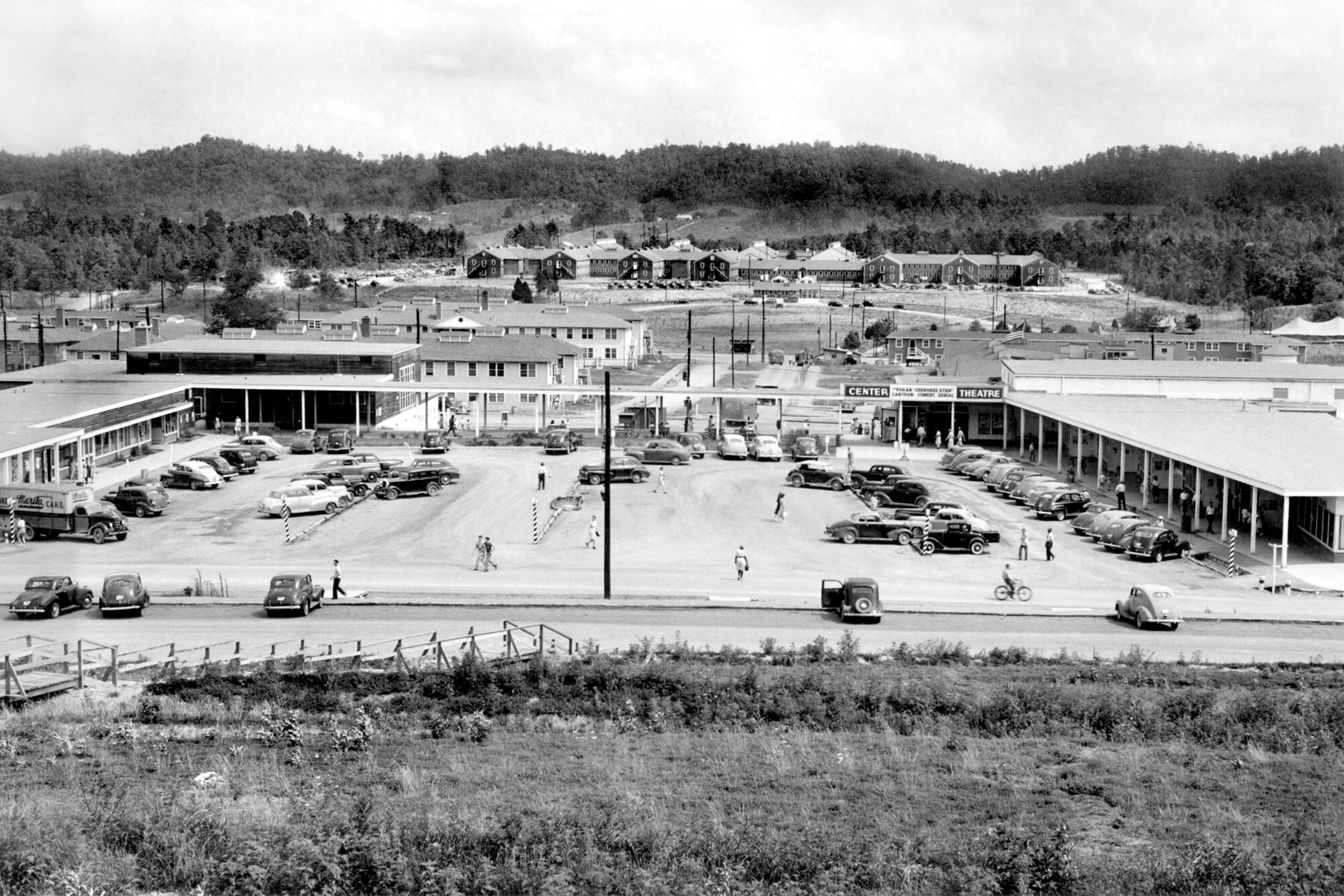 Elevated 1945 photo of a strip mall with parking lot at center and people walking throughout