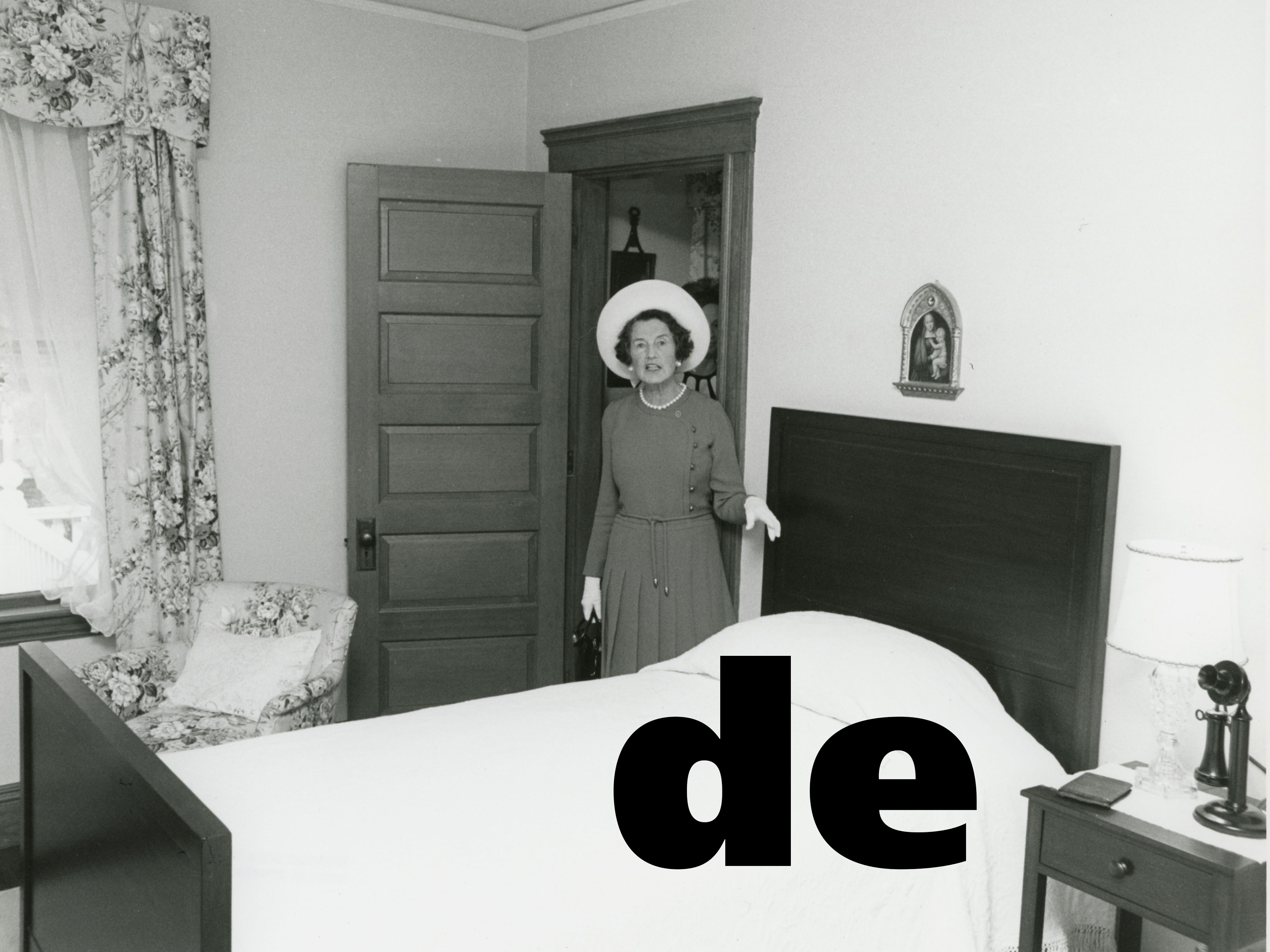 black and white photograph of woman in large white hat standing next to twin bed