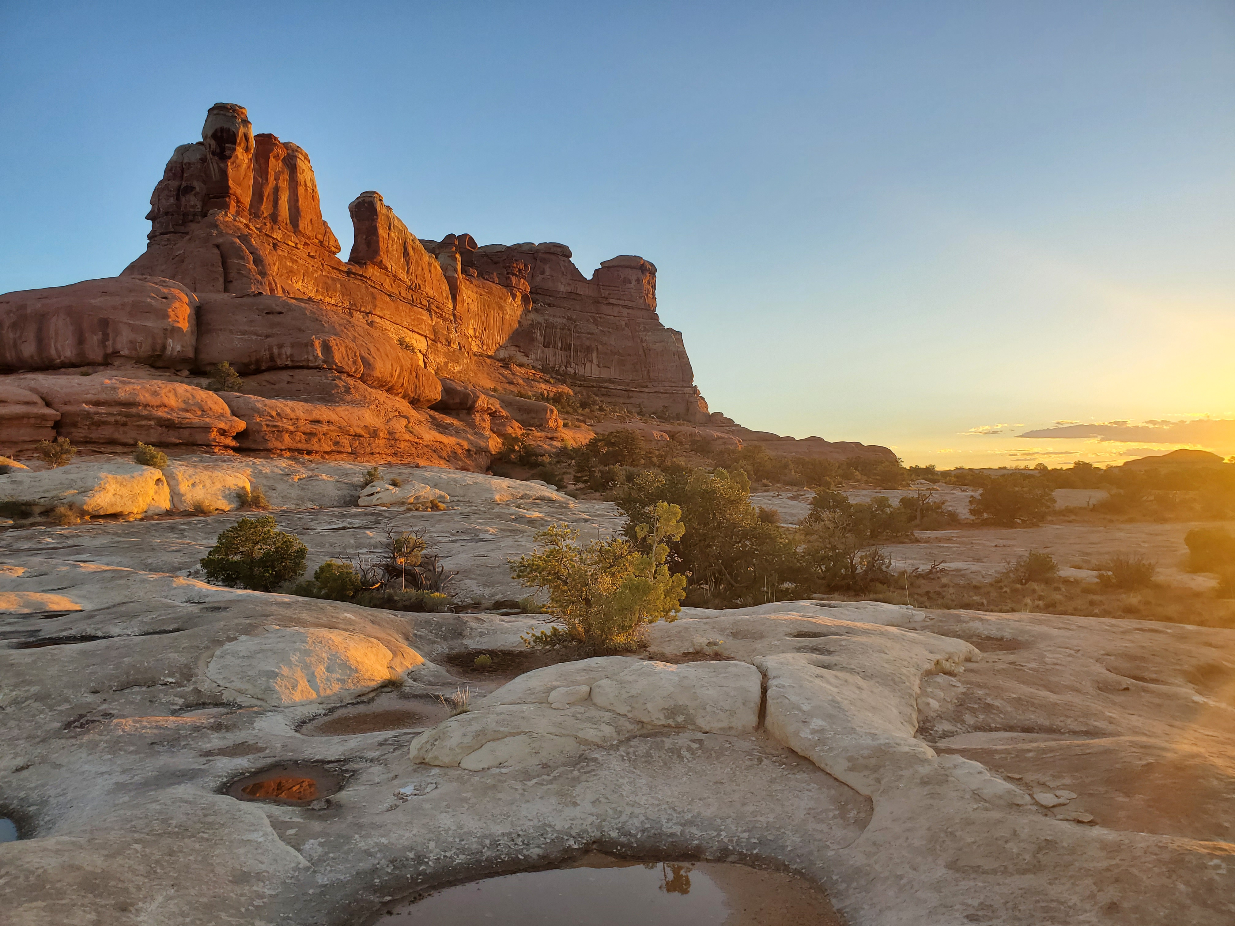 The sun sinks below the horizon behind a vast landscape of slickrock and desert shrubbery.