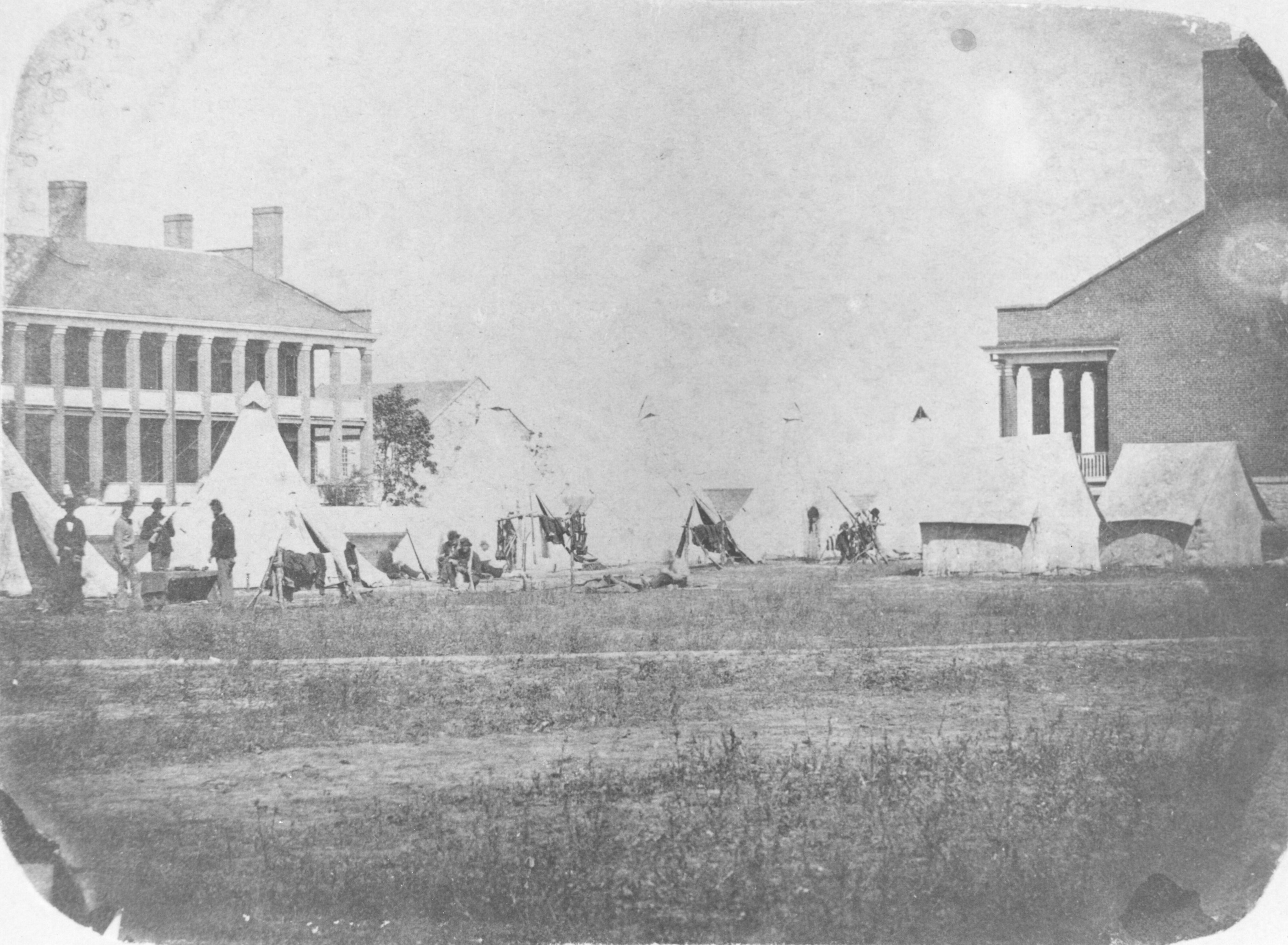 Faded black and white photo soldiers and tents in the foreground and buildings in the background.