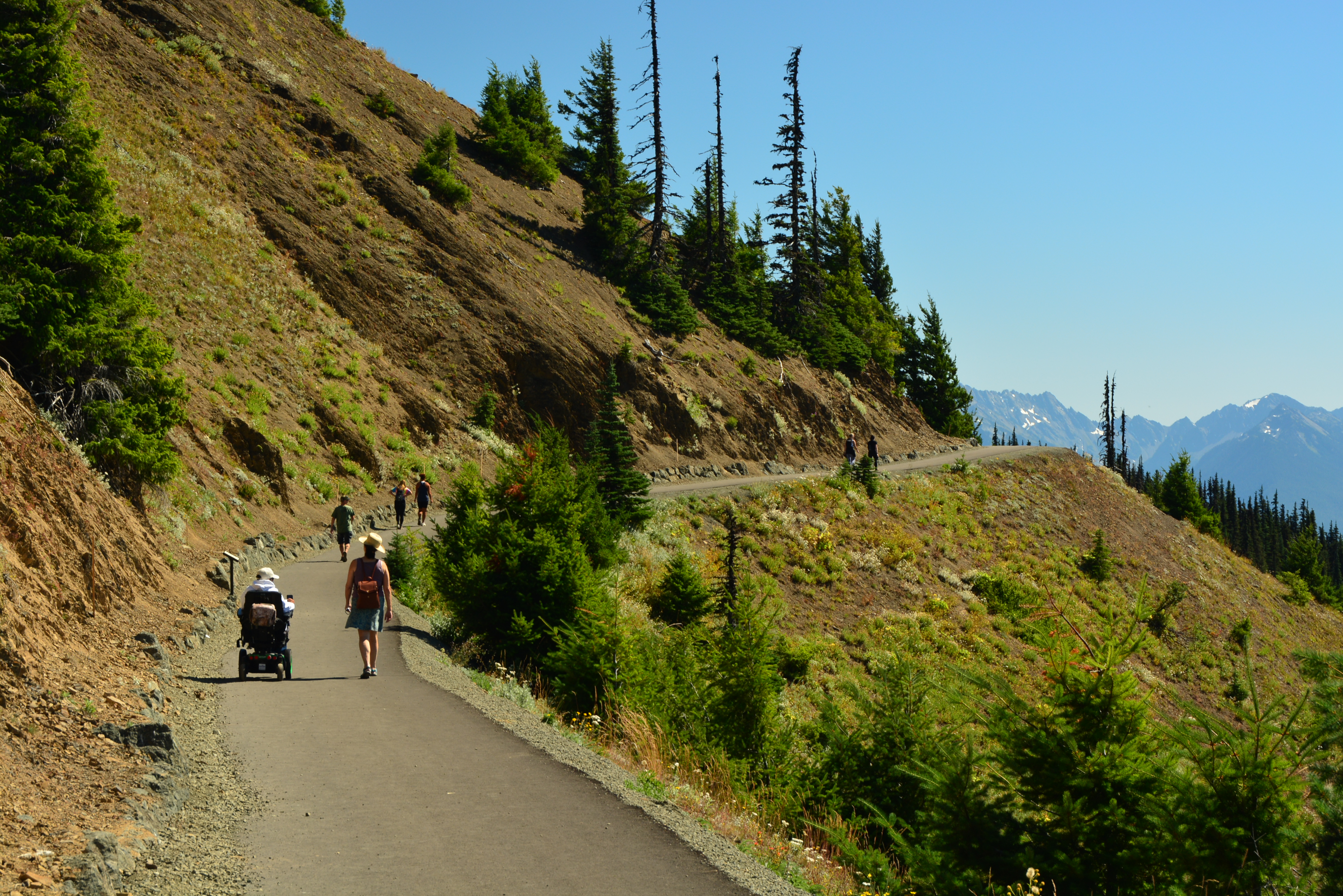 A hiker and wheelchair user on a paved trail cut into the side of a mountain