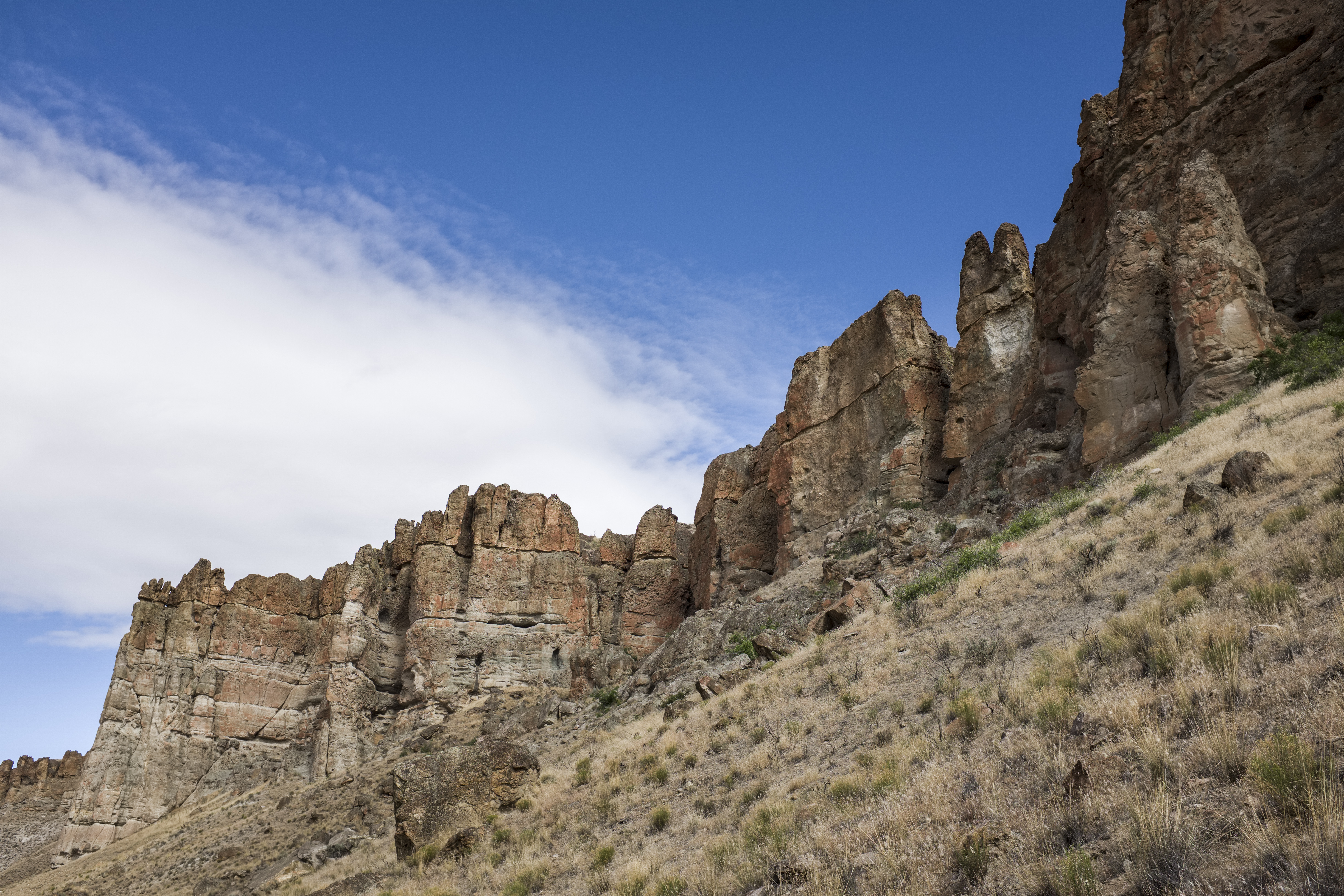 Tan, towering mudflow cliff formations with grassy slopes at its base on a sunny day