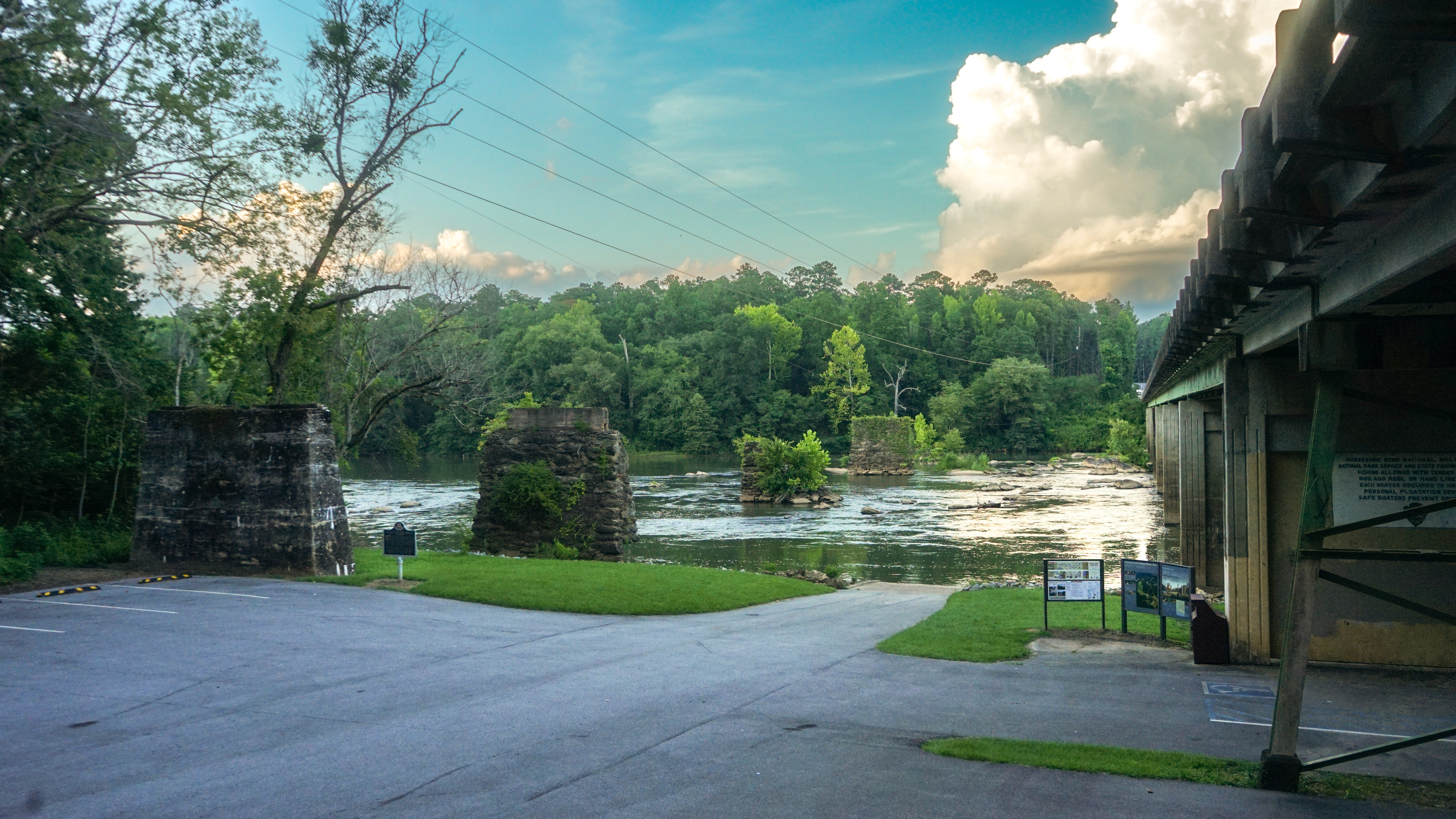 The image displays a small parking lot next to a flowing river and a large roadway bridge.