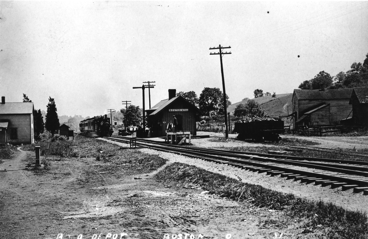 Black-and-white photo of a train approaching a station; a sign on the station reads "Boston Mill".