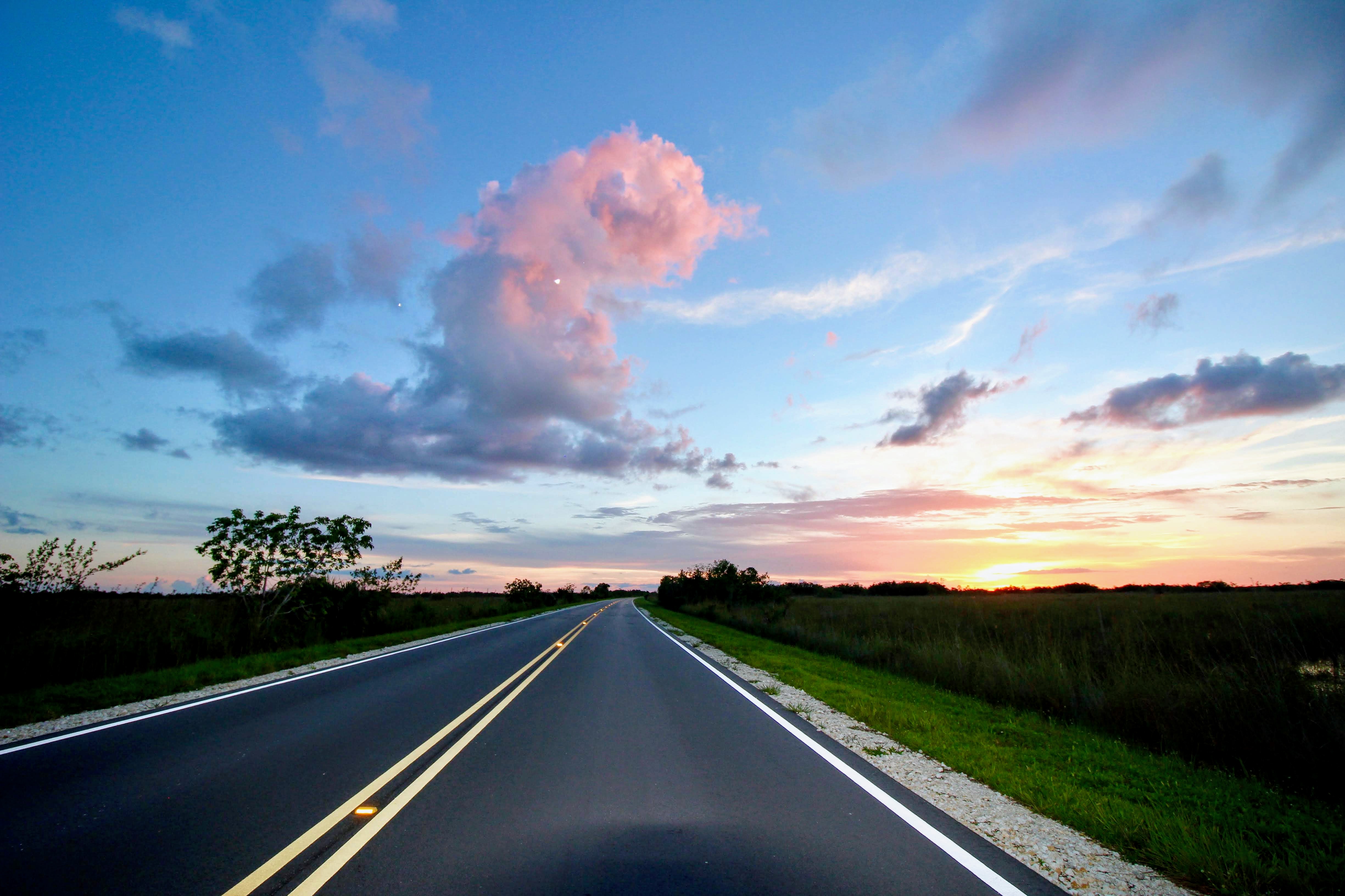 A paved, double striped road travels into the distance under a cloudy, blue and purple sunset sky