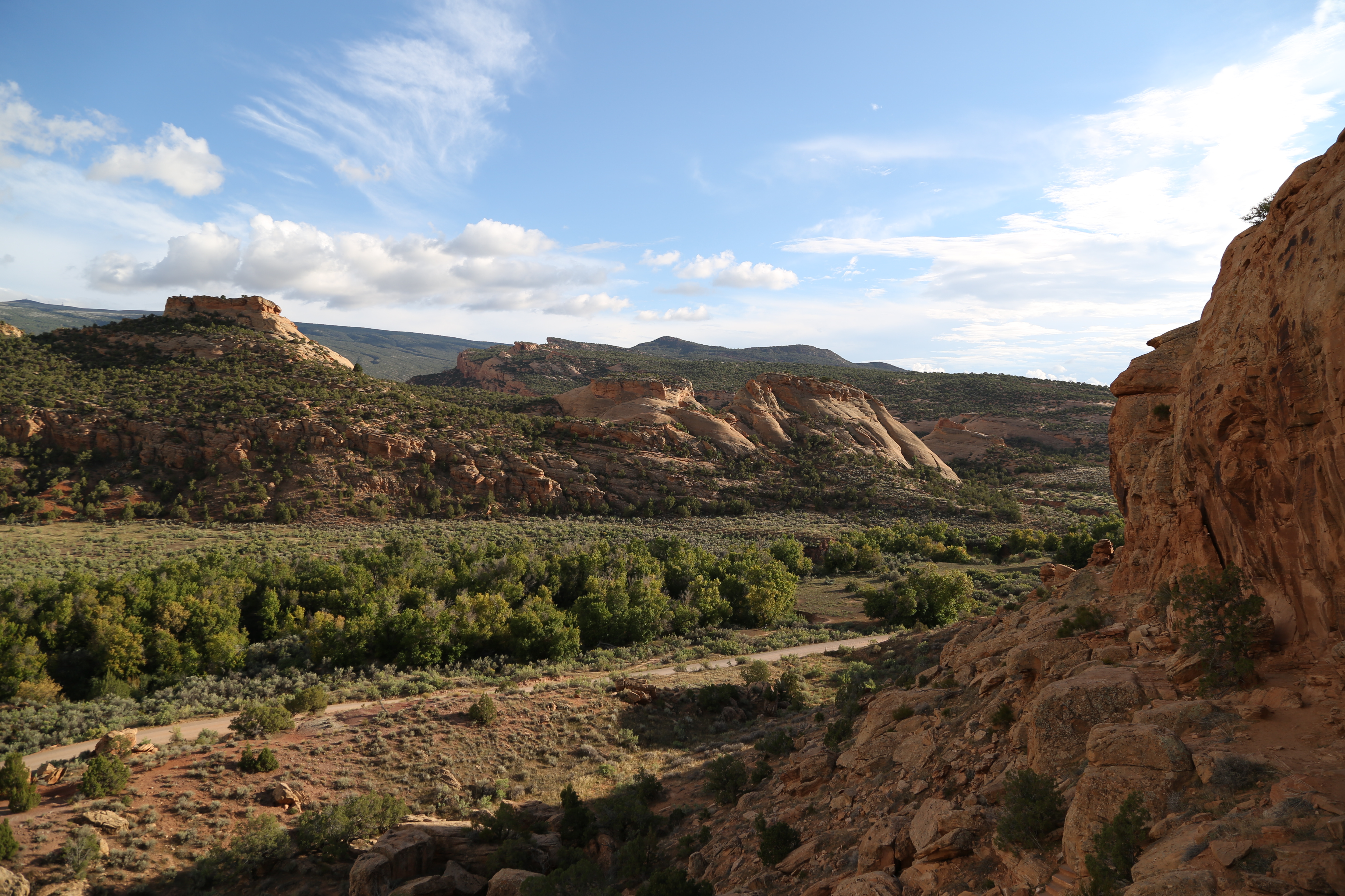 Orange colored sandstone bluffs rising above desert shrubs under a partly cloudy blue sky.