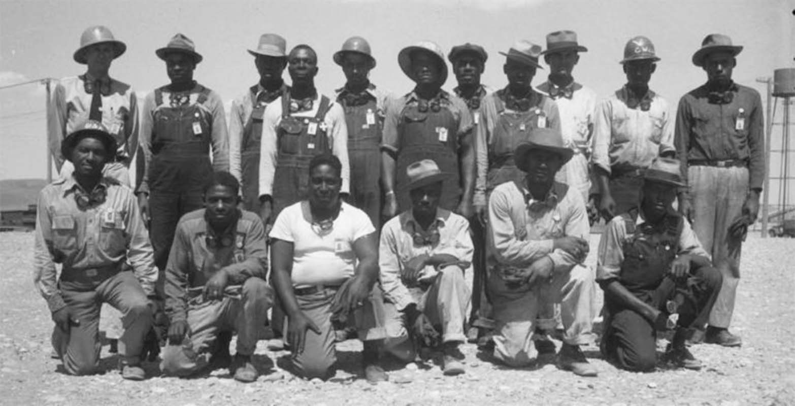 A group of 18 men in work clothes and protective hats pose for a picture. Six men kneel in the front