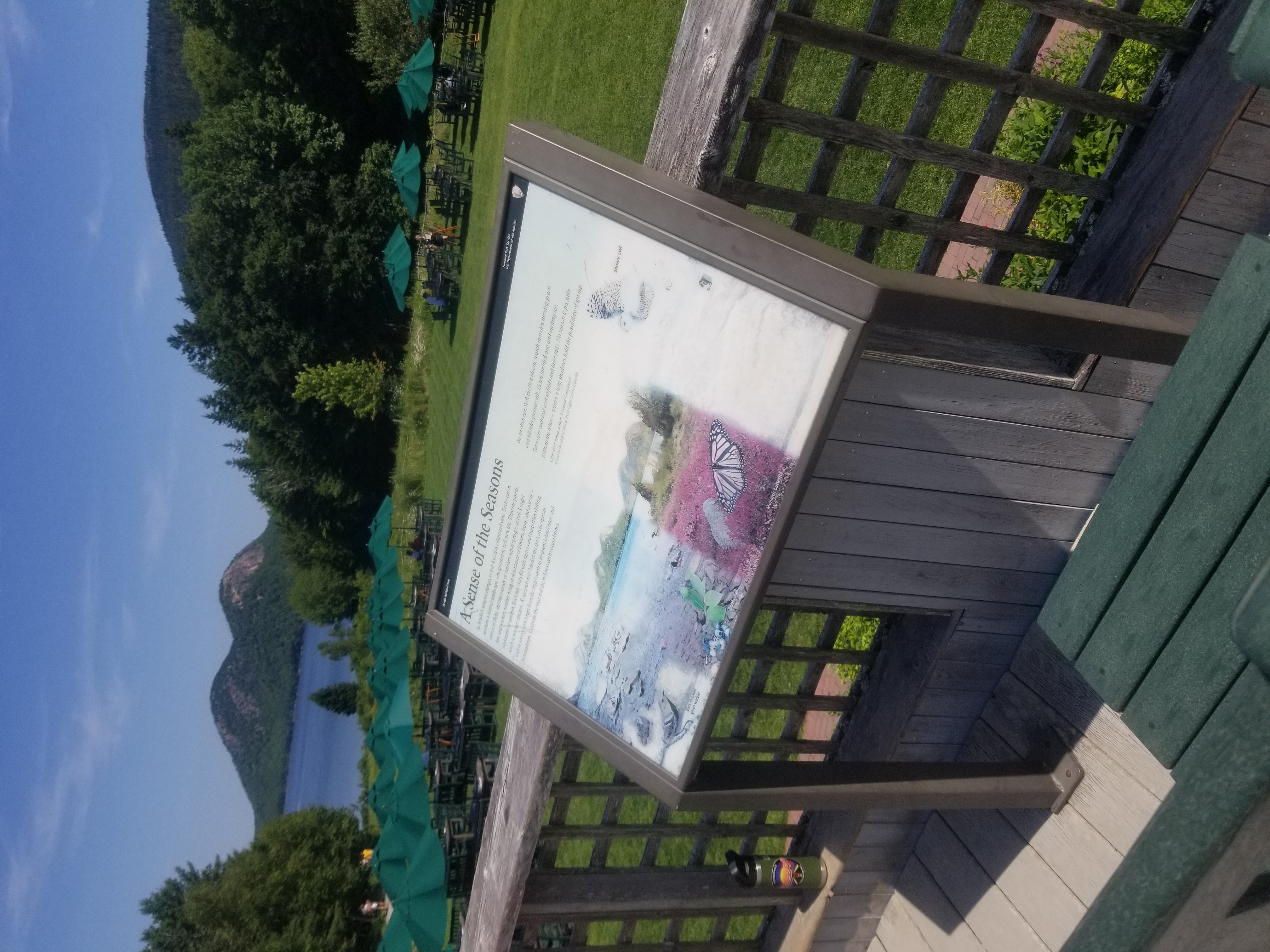 A wayside placed on a deck overlooking a lawn with restaurant tables and sun umbrellas.