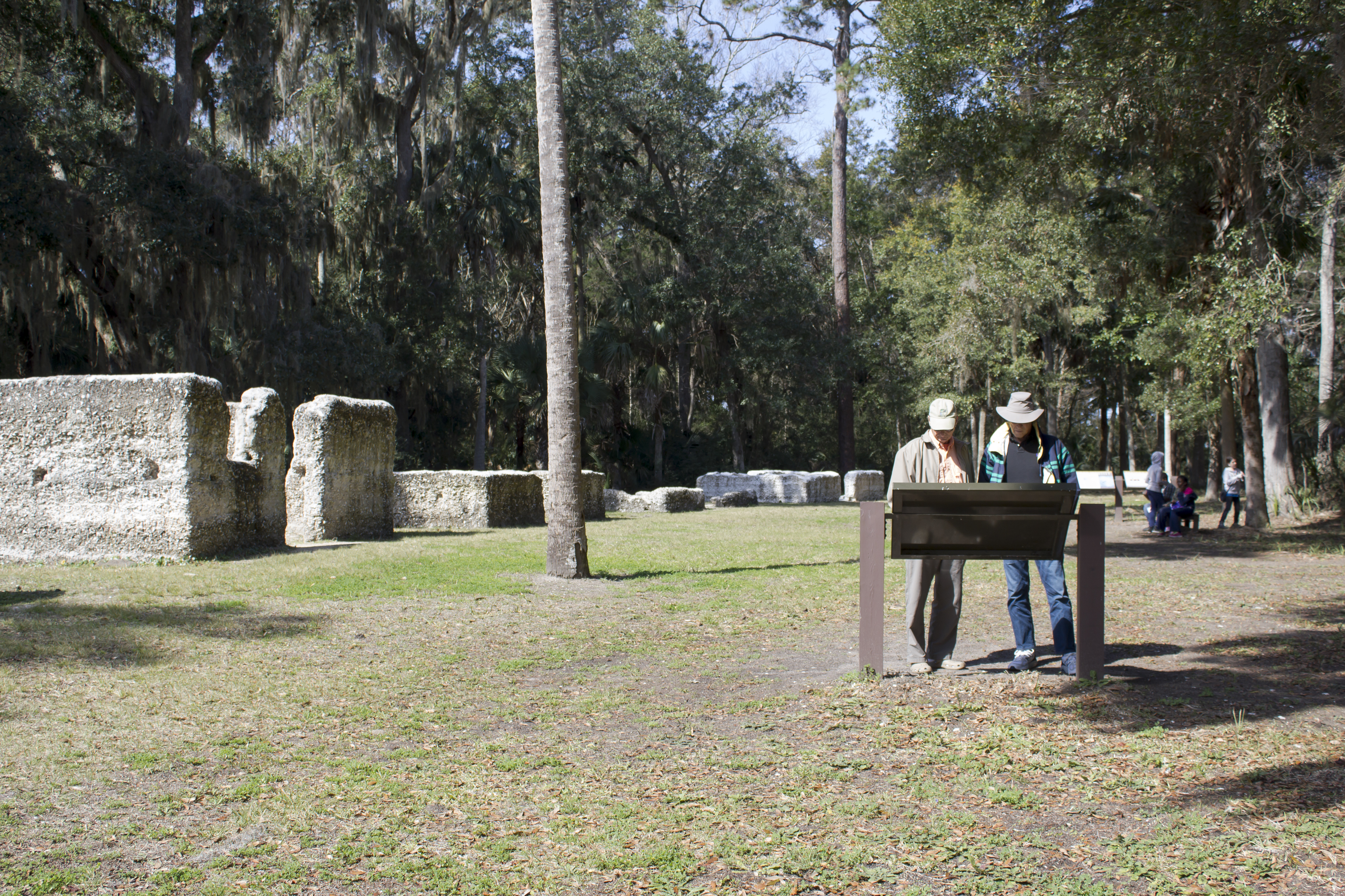 two visitors read an outdoor exhibit in front of white roofless tabby buildings