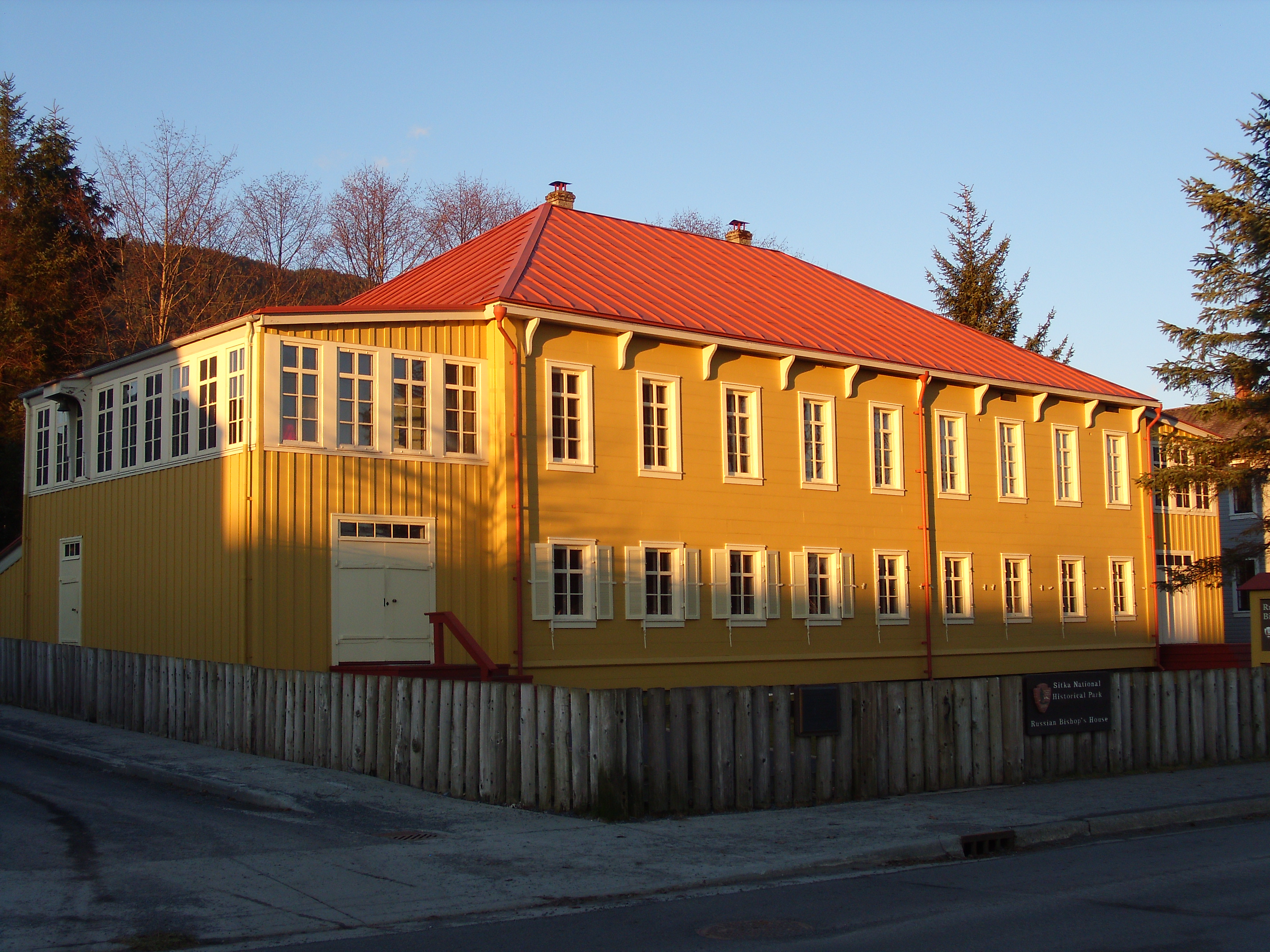 A large, yellow, two story building with a terra cotta roof, surrounded by fence and sidewalk.