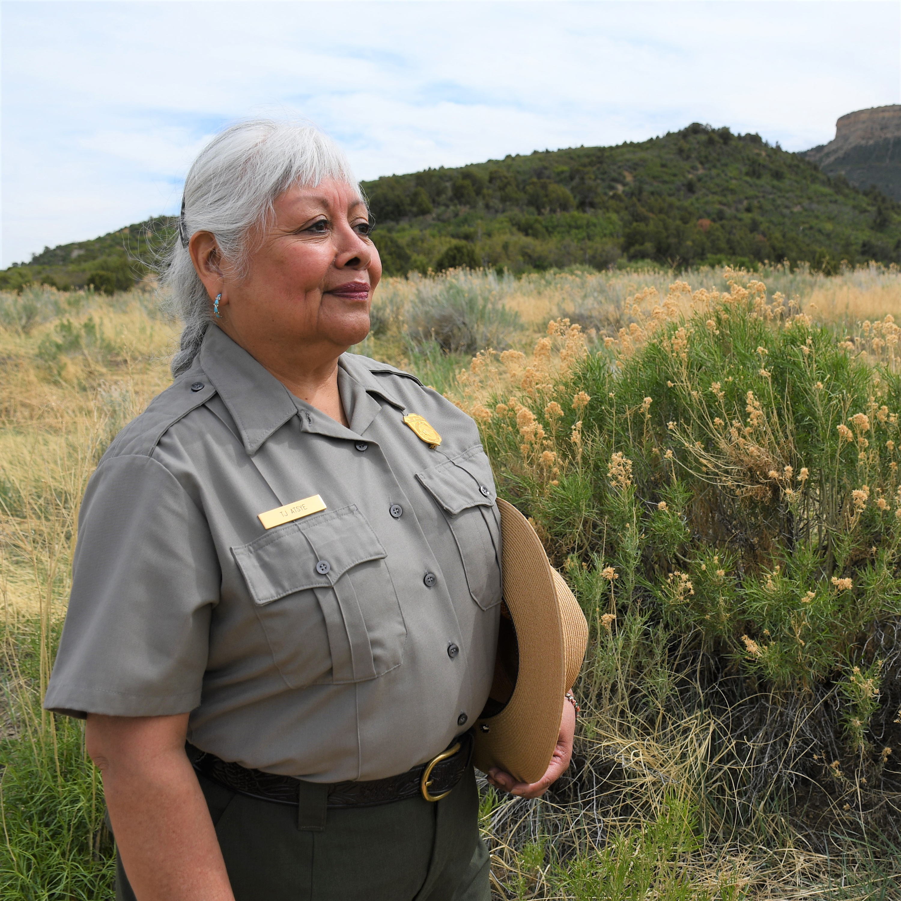 A Pueblo woman in ranger uniform with long gray hair stands in front of rabbitbrush and foothills