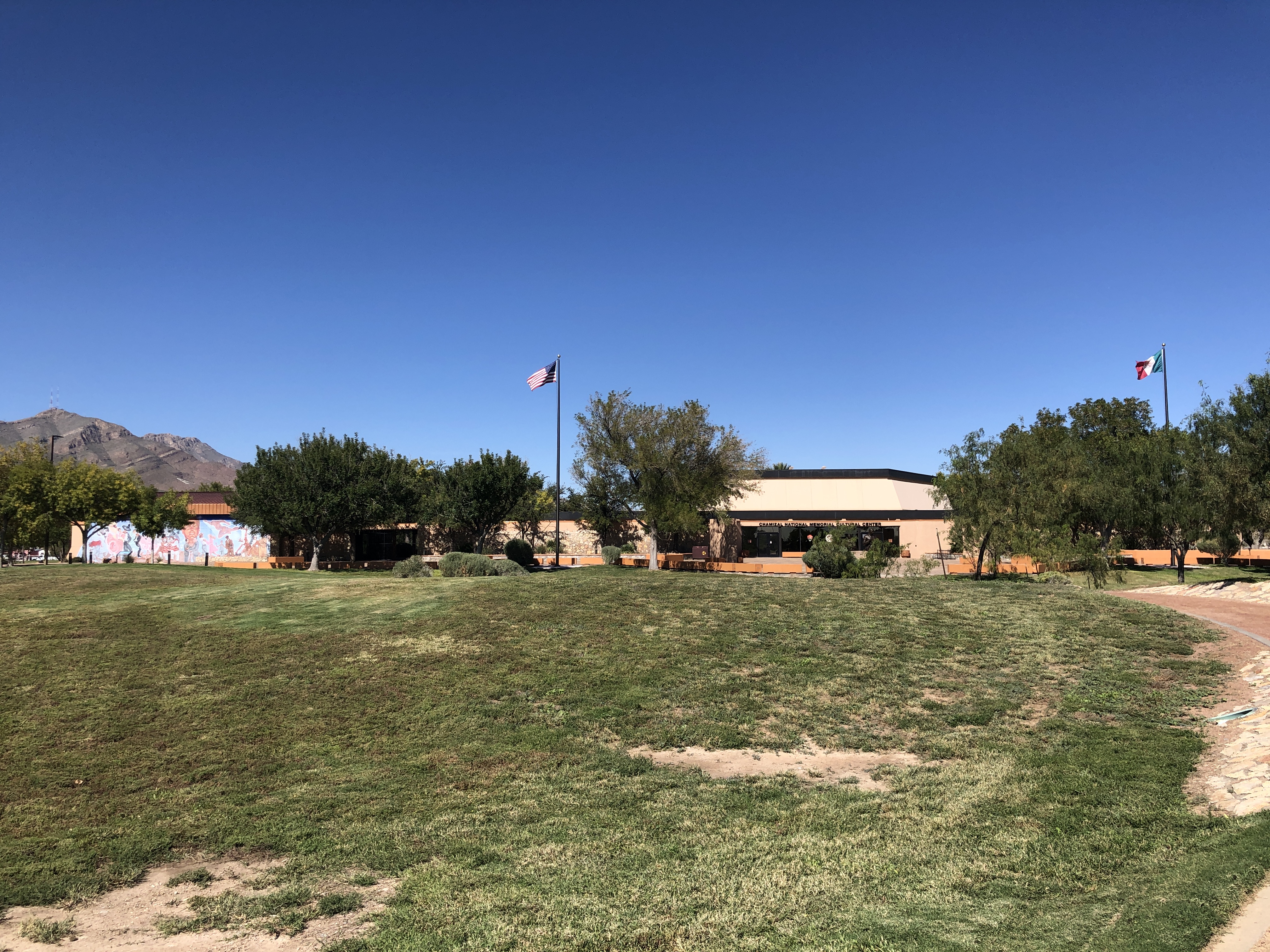 A tan building, two flag poles, a section of mural, and mountaintop across expanse of grass and sky