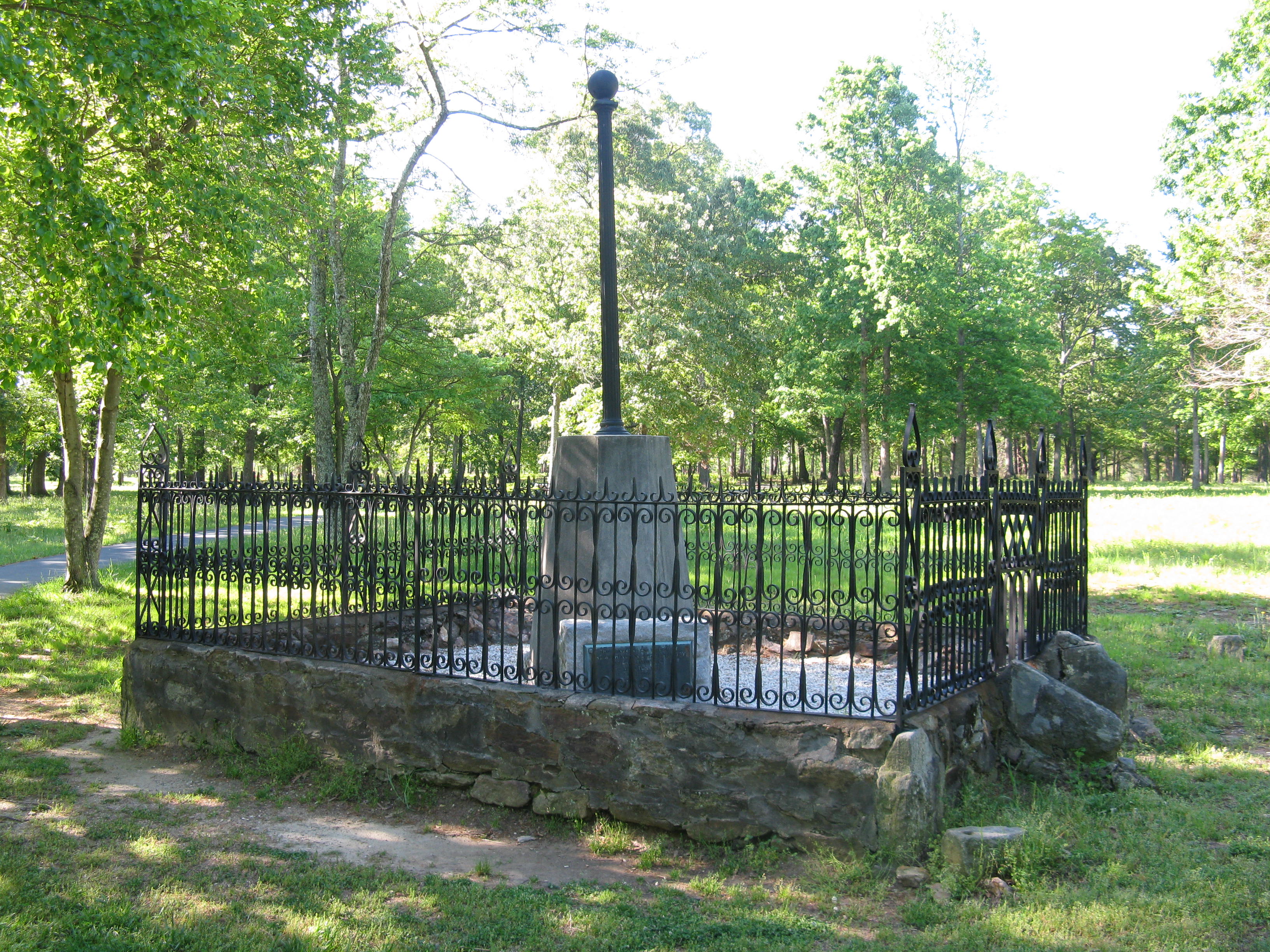 A fence made of a rock base and iron stakes surround a monument made of concrete and flagpole.