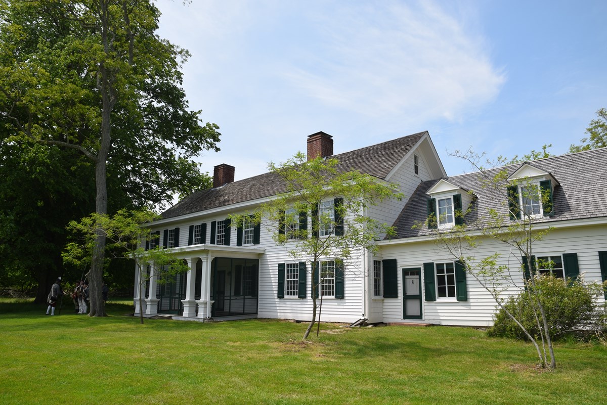 A white manor house with green trim is surrounded by trees and sky.