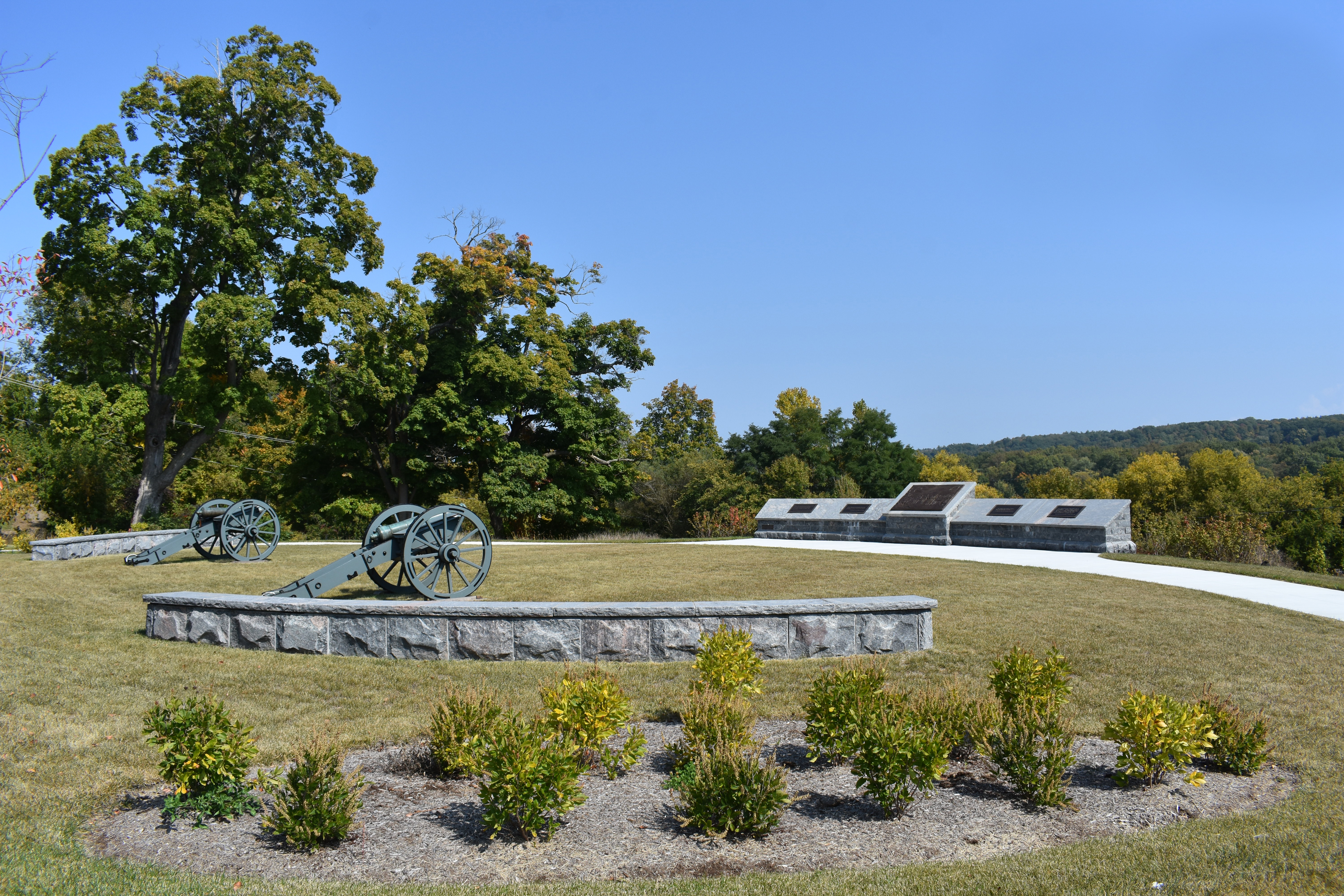 Two cannons on a grassy lawn near a short stone informational wall sign with bronze plaques on it.